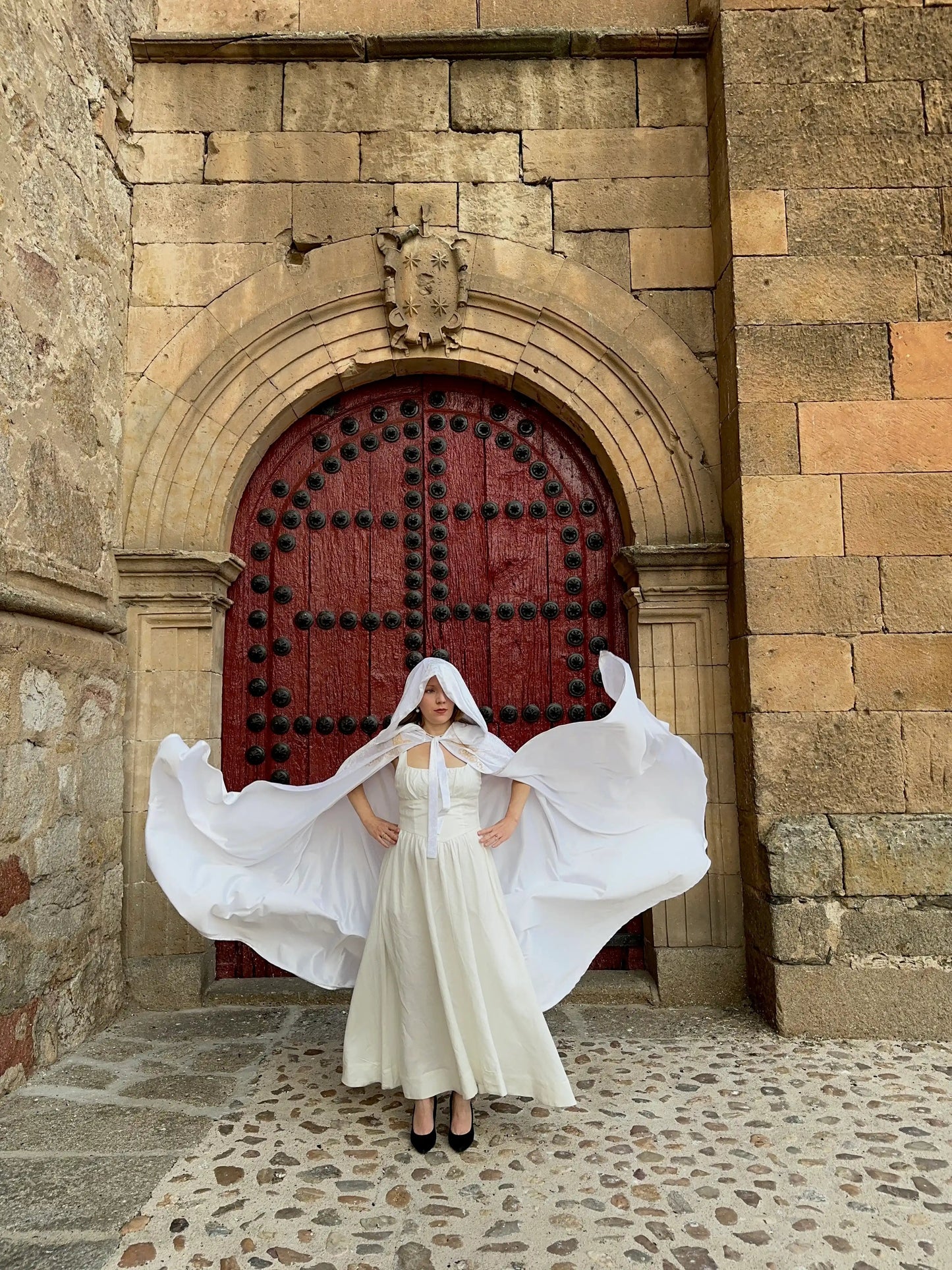 Woman in a white dress with a flowing cape standing in front of a large red door on a stone building.