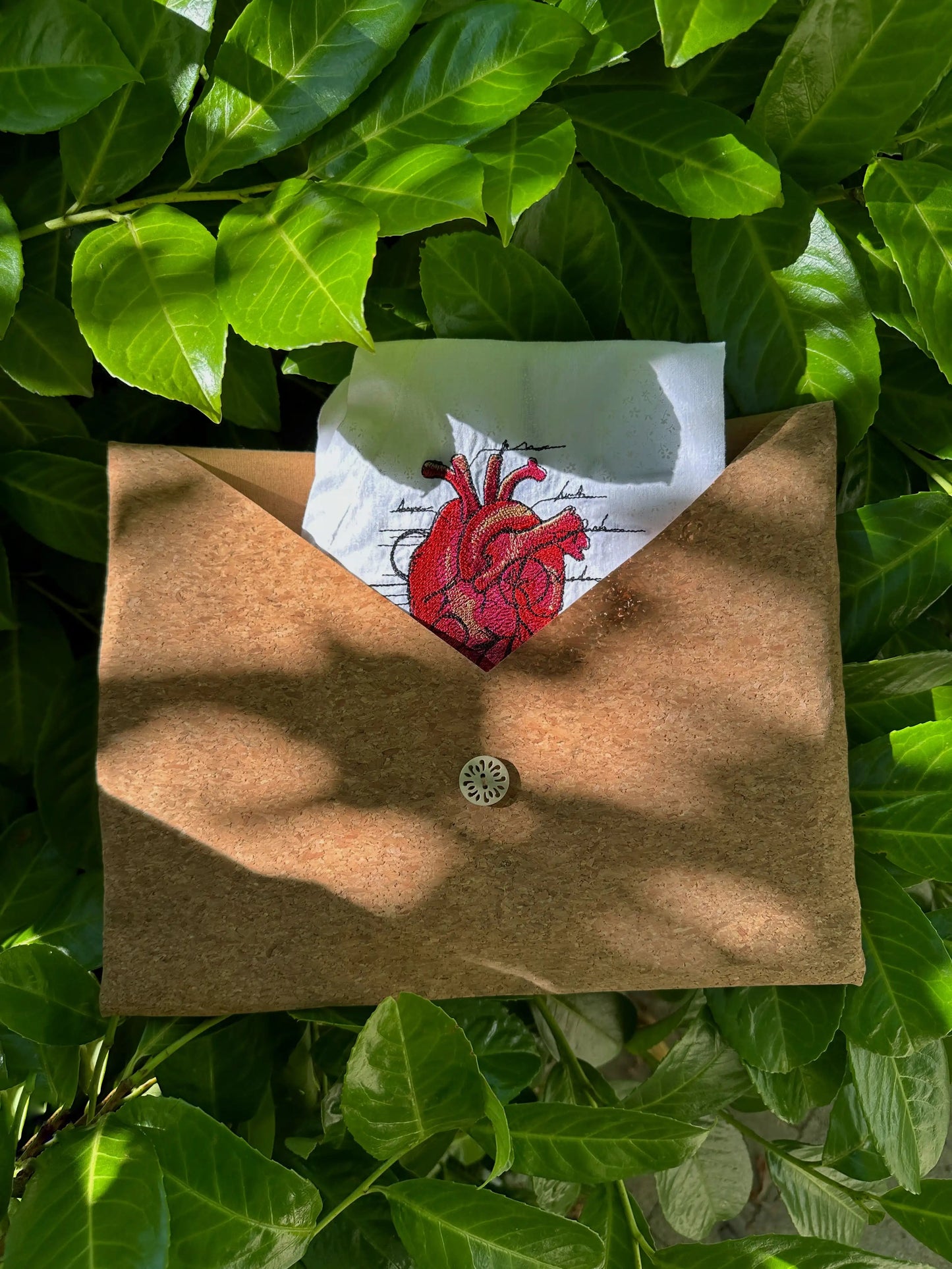 Brown envelope with a red embroidered heart design on a white card, placed on green leaves.