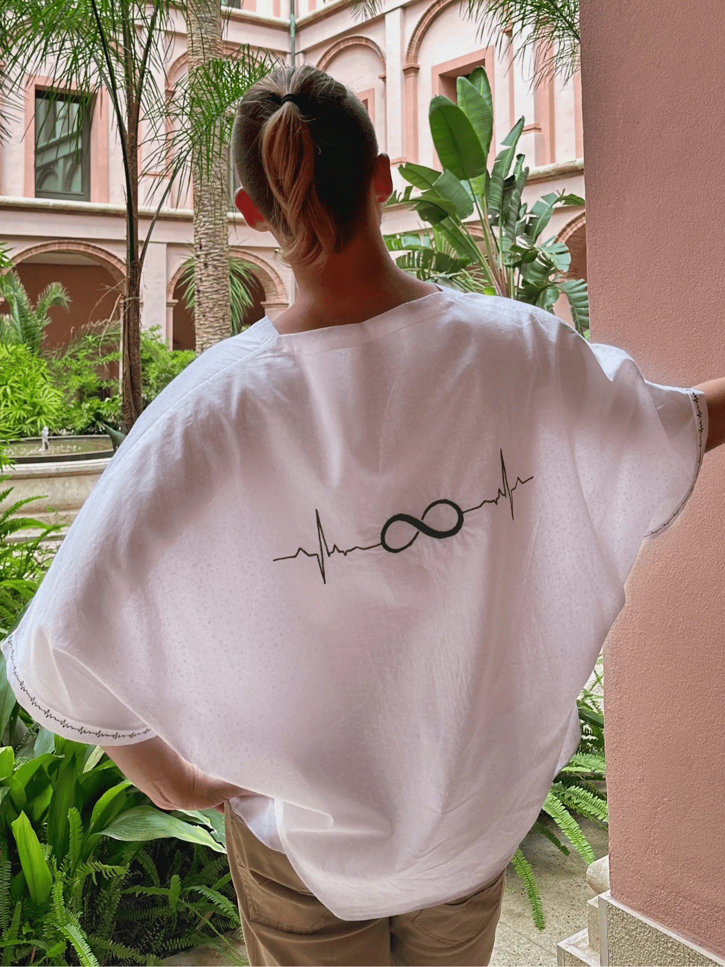 Person wearing a white shirt with a design, standing in a courtyard with pink walls and greenery.