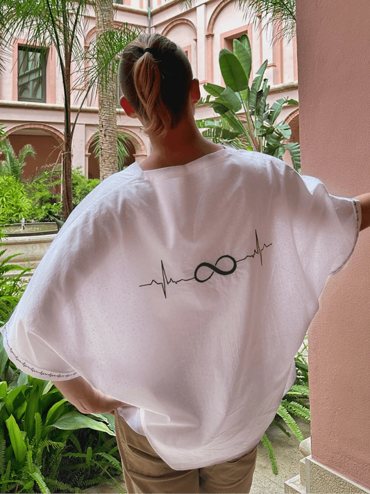 Person wearing a white shirt with a design, standing in a courtyard with pink walls and greenery.