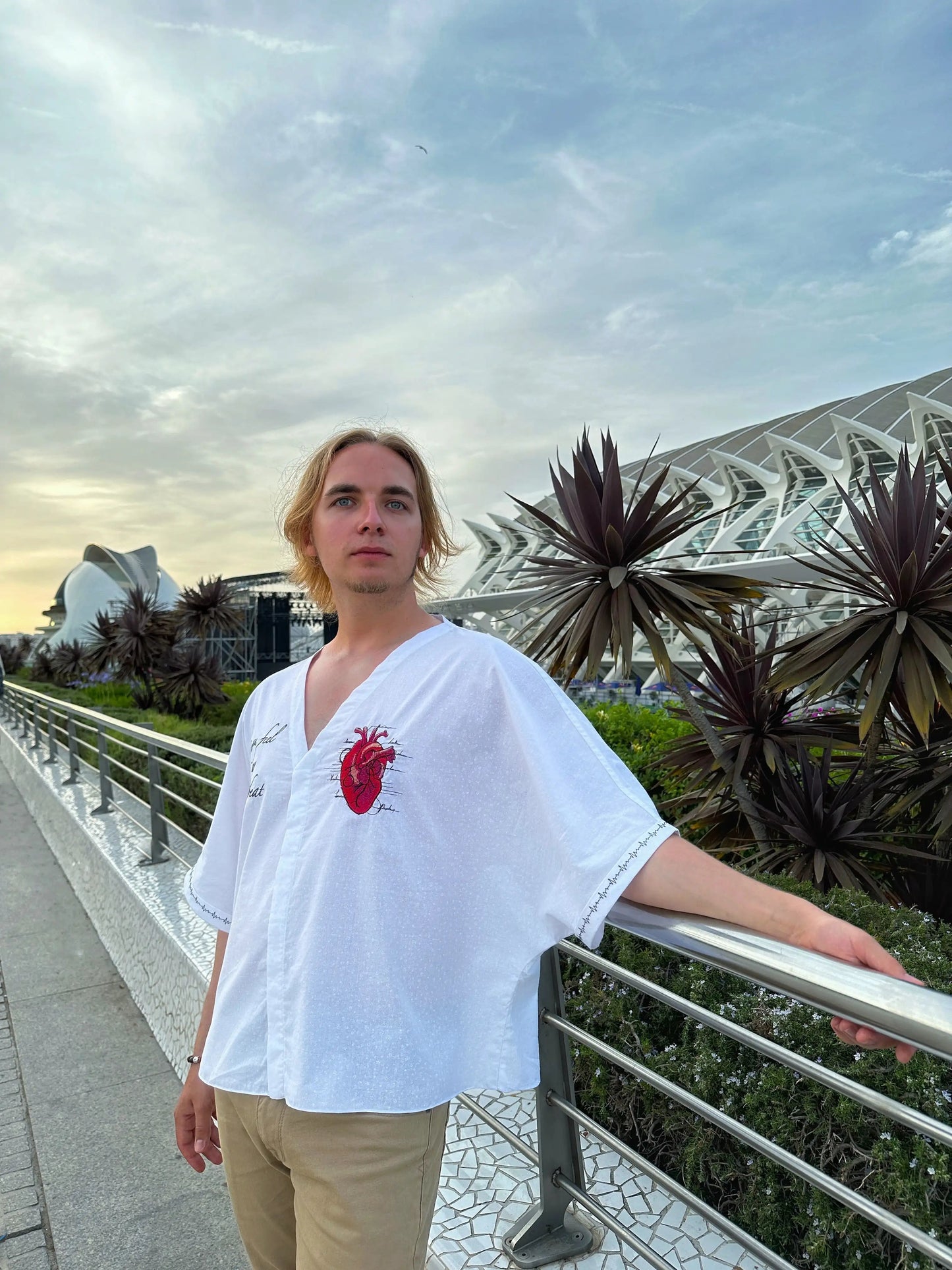 Person wearing a white shirt with a red heart design, standing outdoors with modern architecture and plants in the background.