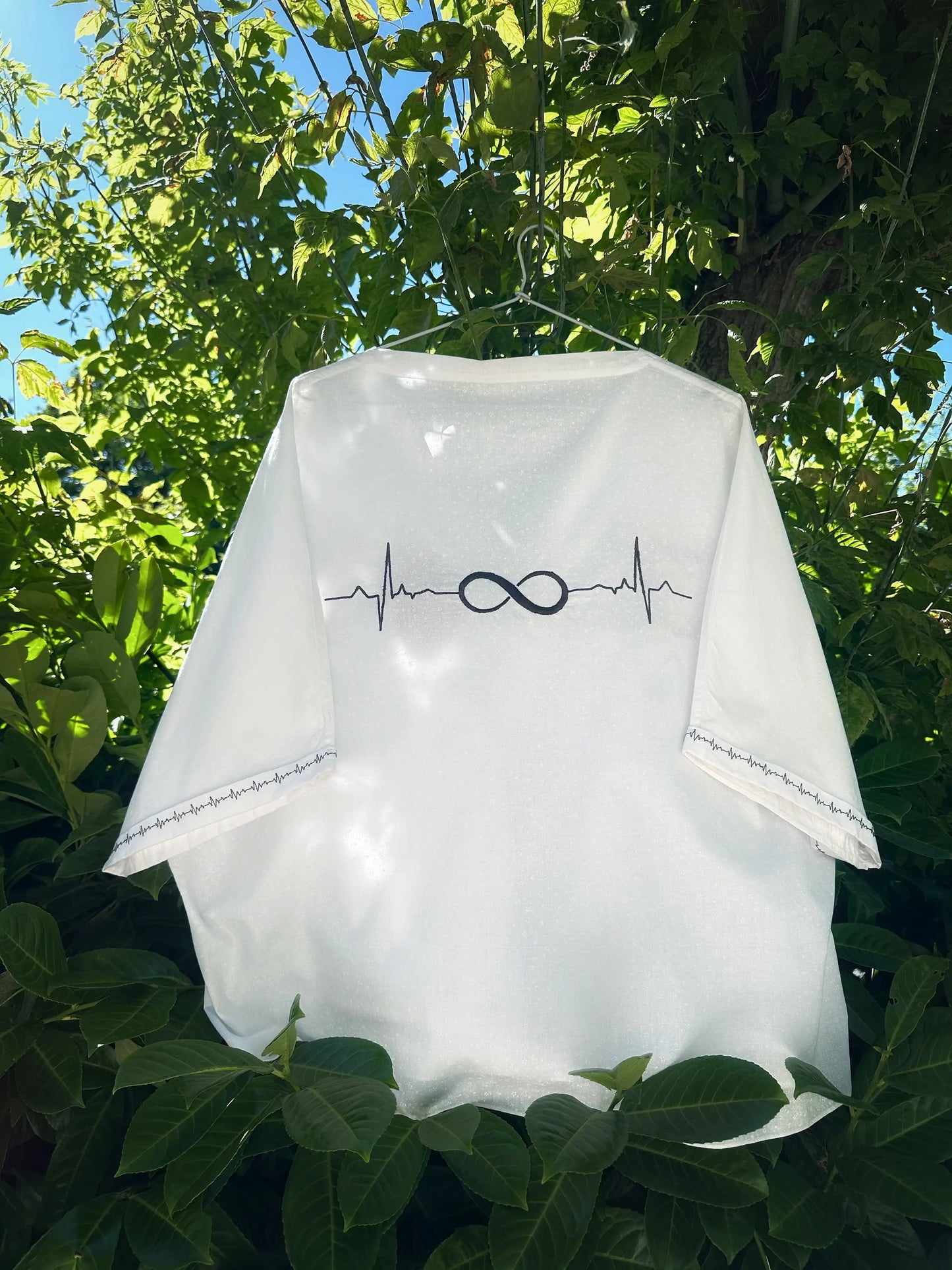 White baby romper with infinity symbol and heartbeat line design, hanging outdoors among green leaves.