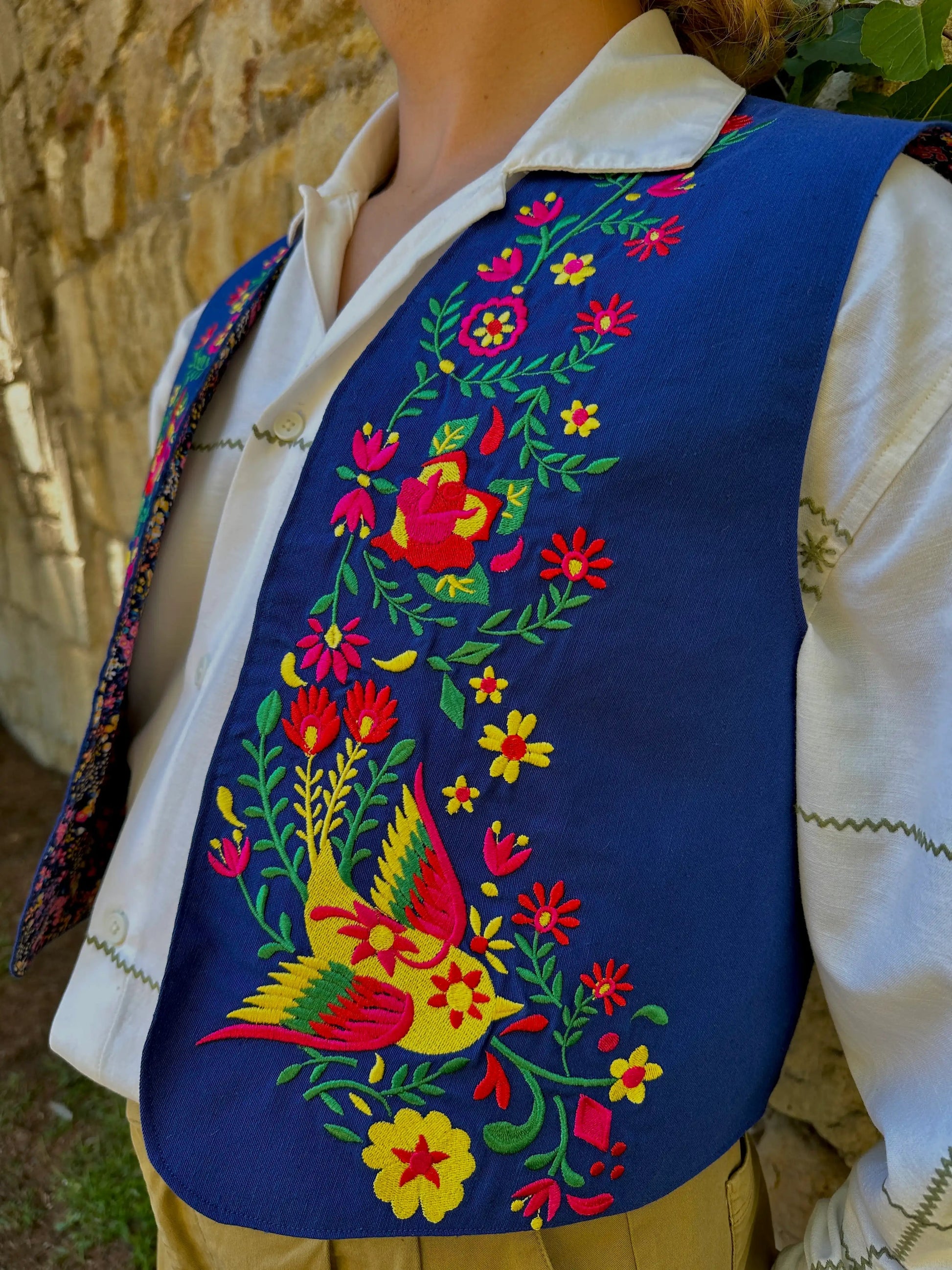 Person wearing a colorful embroidered vest with floral and bird patterns against a stone wall.