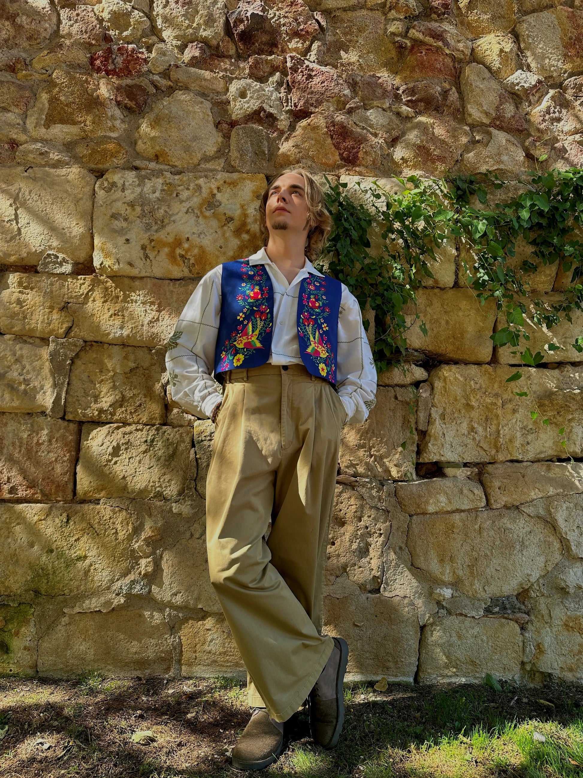Person wearing a colorful vest and beige pants standing against a stone wall.