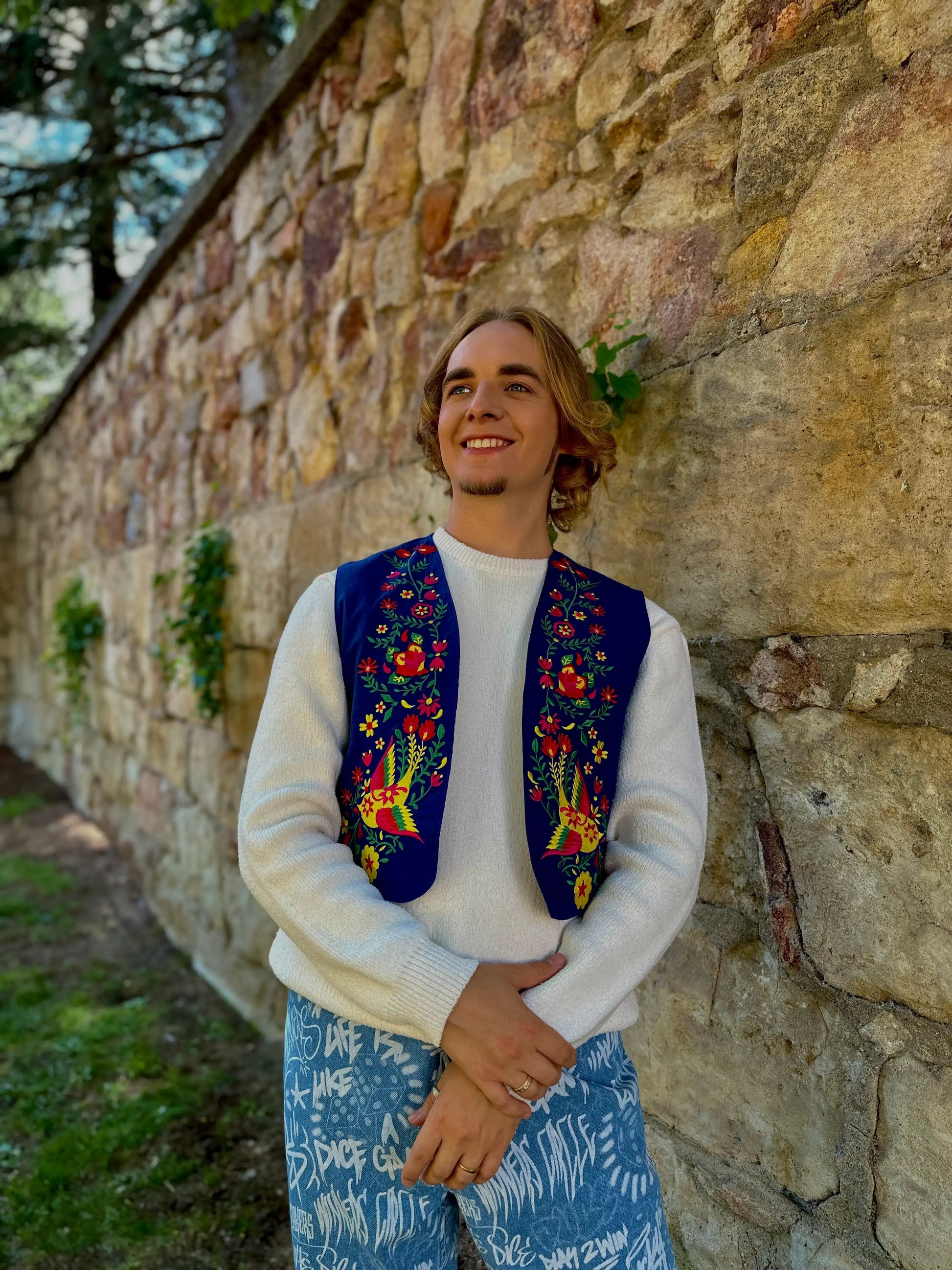 Person wearing a colorful embroidered vest and patterned pants standing against a stone wall.