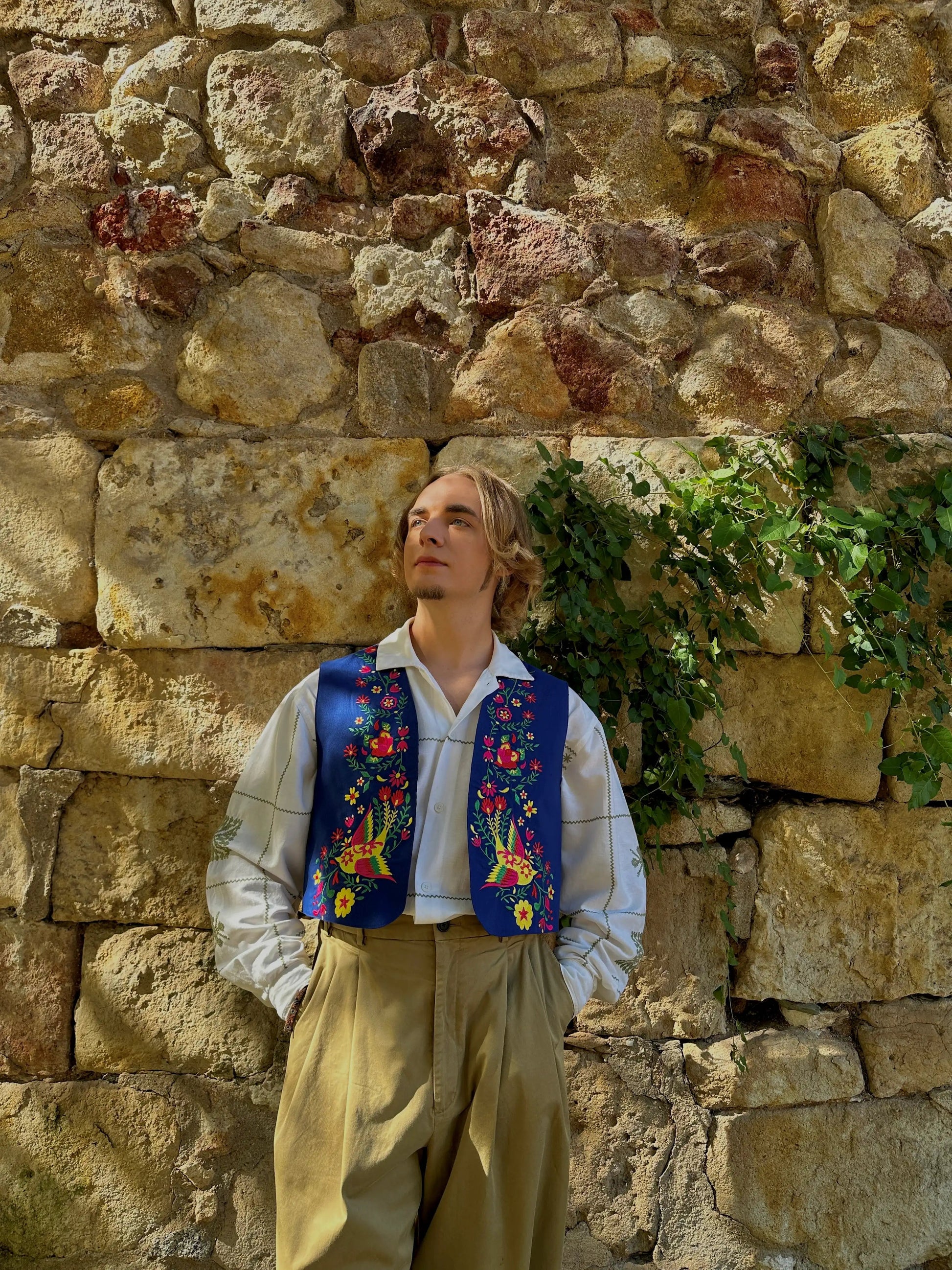 Person wearing a colorful embroidered vest against a stone wall