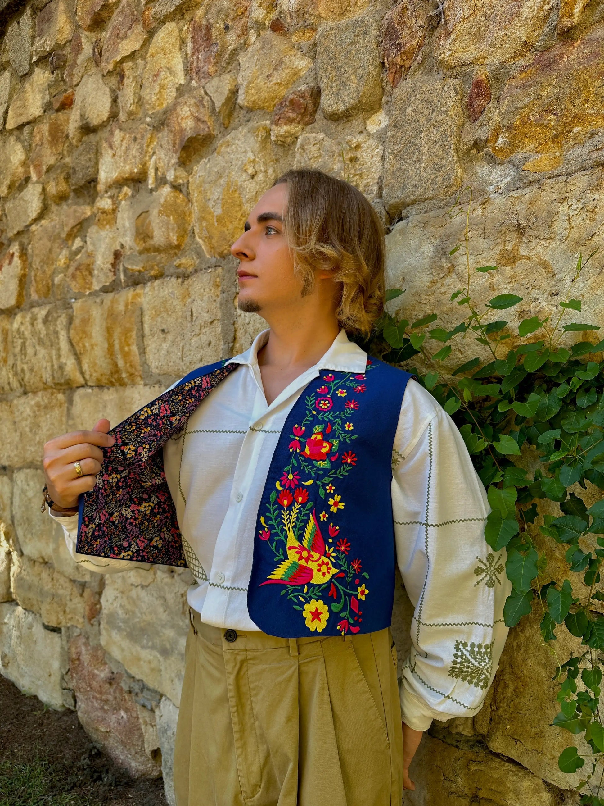 Man wearing a colorful embroidered vest against a stone wall