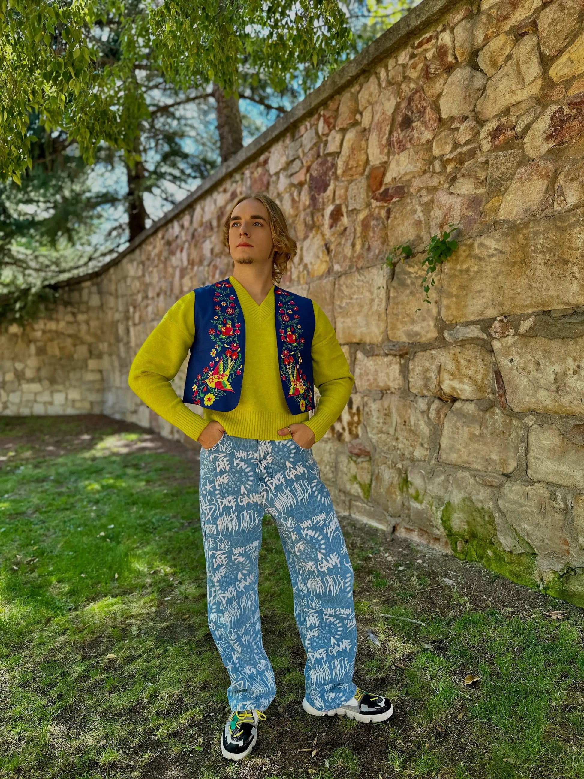 Person wearing a colorful vest over a yellow shirt with blue patterned pants, standing against a stone wall.