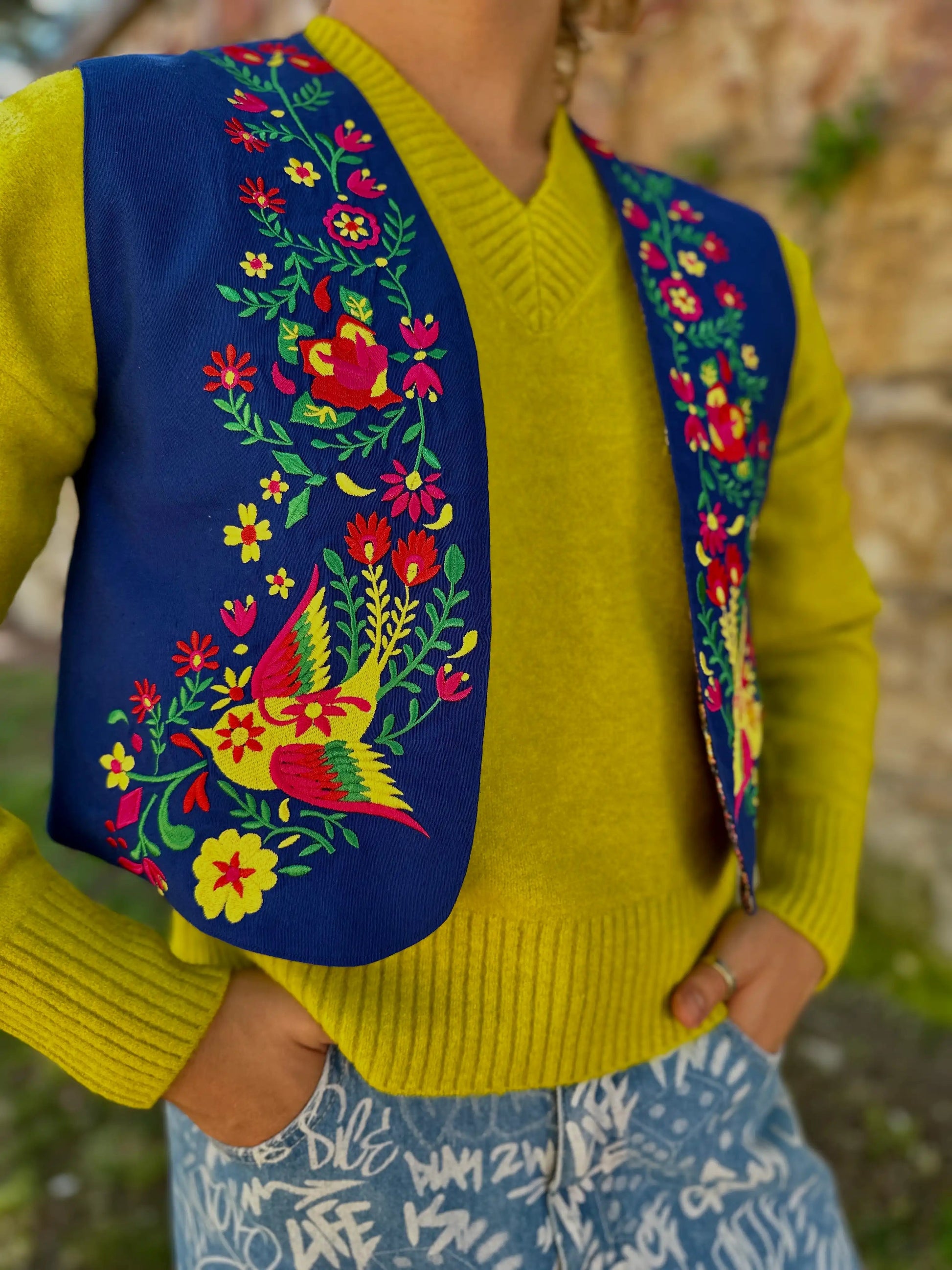Person wearing a bright yellow sweater with a colorful embroidered blue vest over it, standing outdoors.
