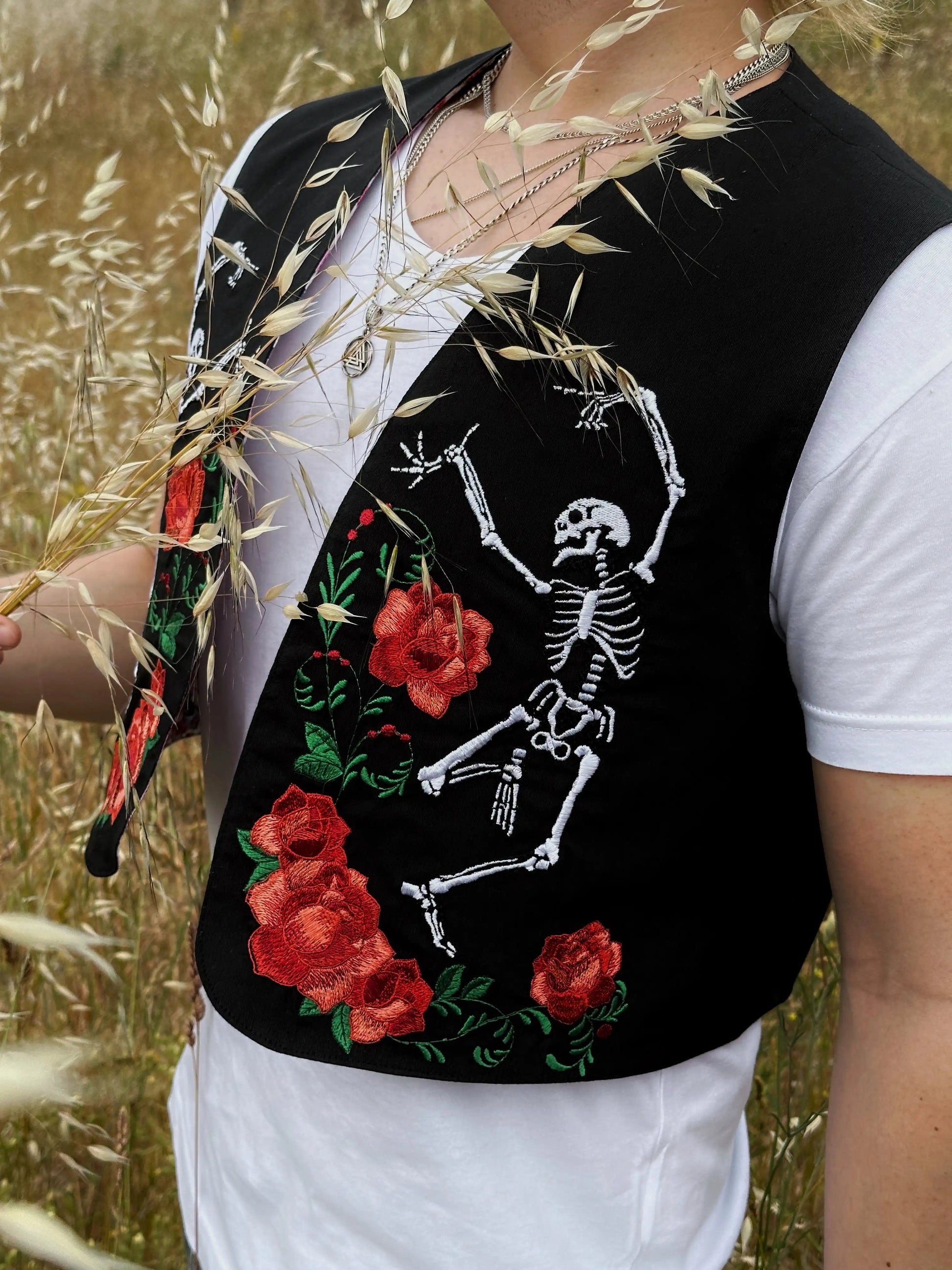 Person wearing a black vest with skeleton and rose embroidery in a field.