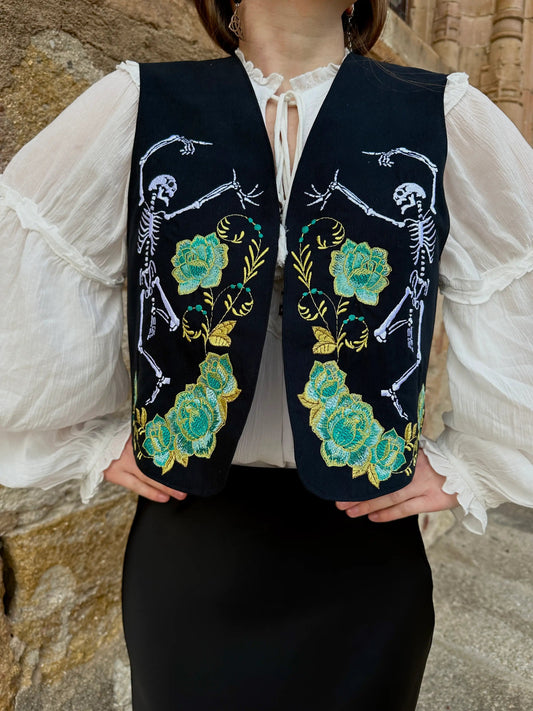 Person wearing a black vest with floral and skeleton designs against a stone wall.
