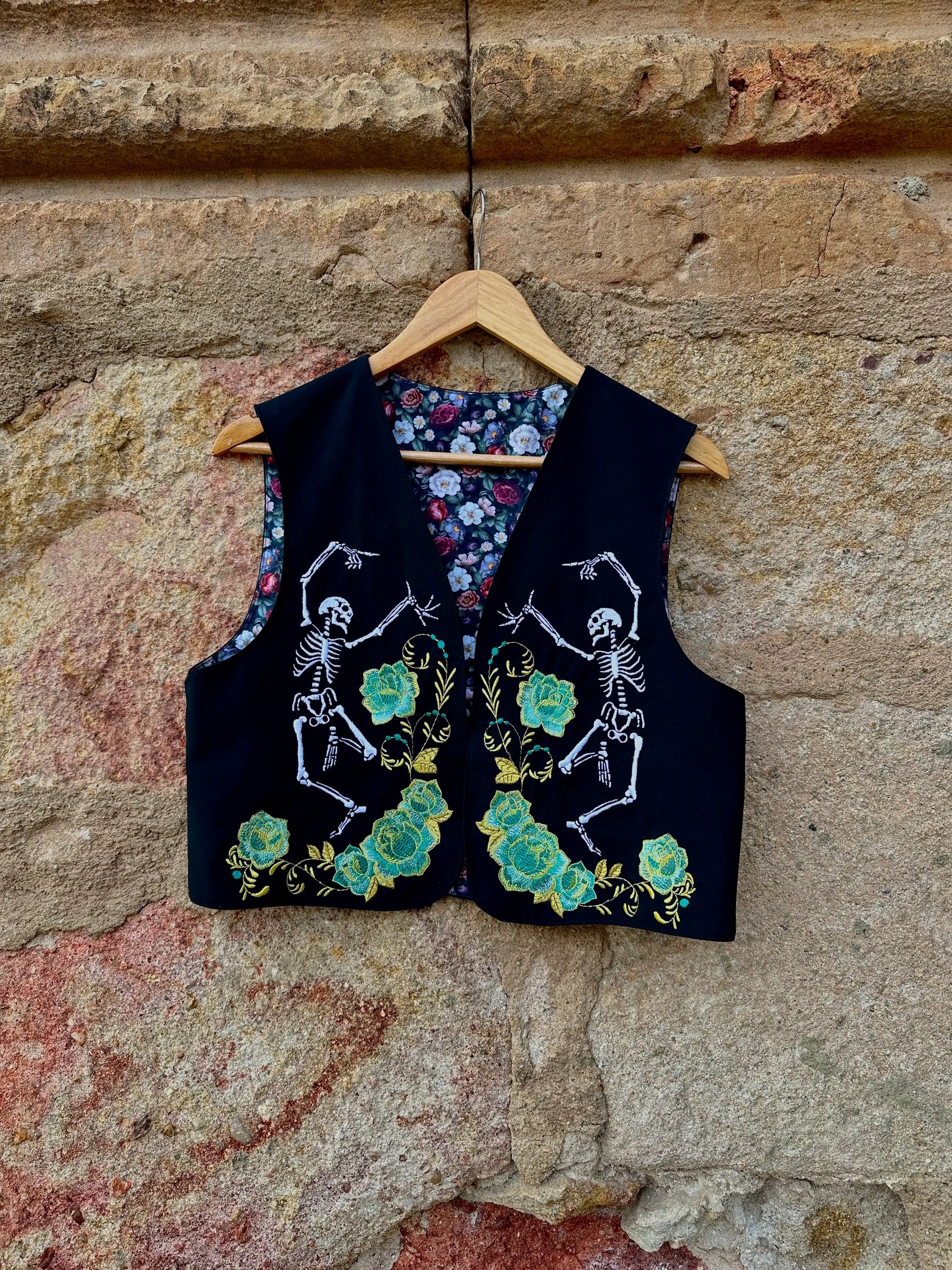Black vest with skeleton and floral design on a wooden hanger against a stone wall.
