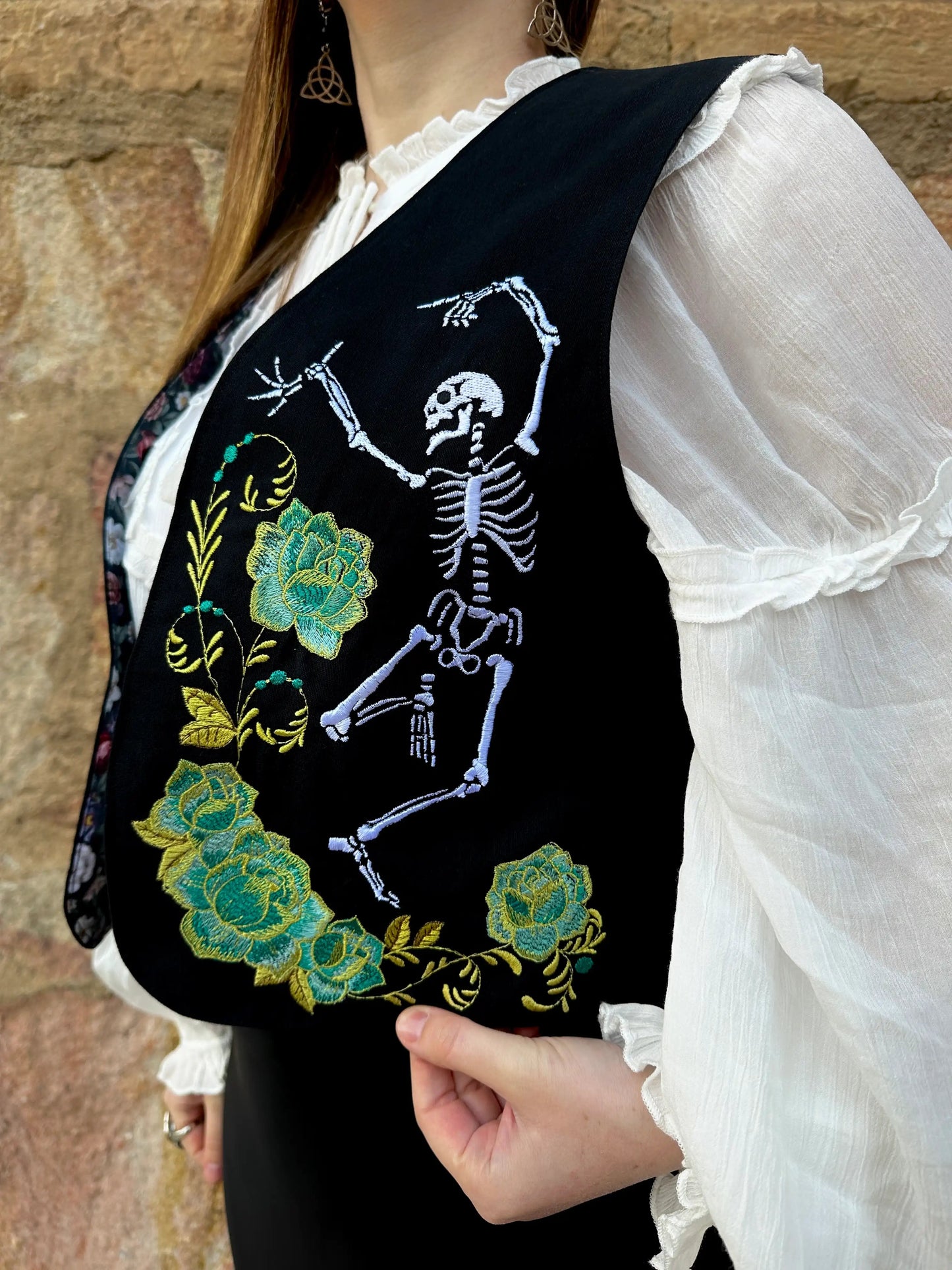 Black vest with skeleton and floral design worn by a person against a stone wall.