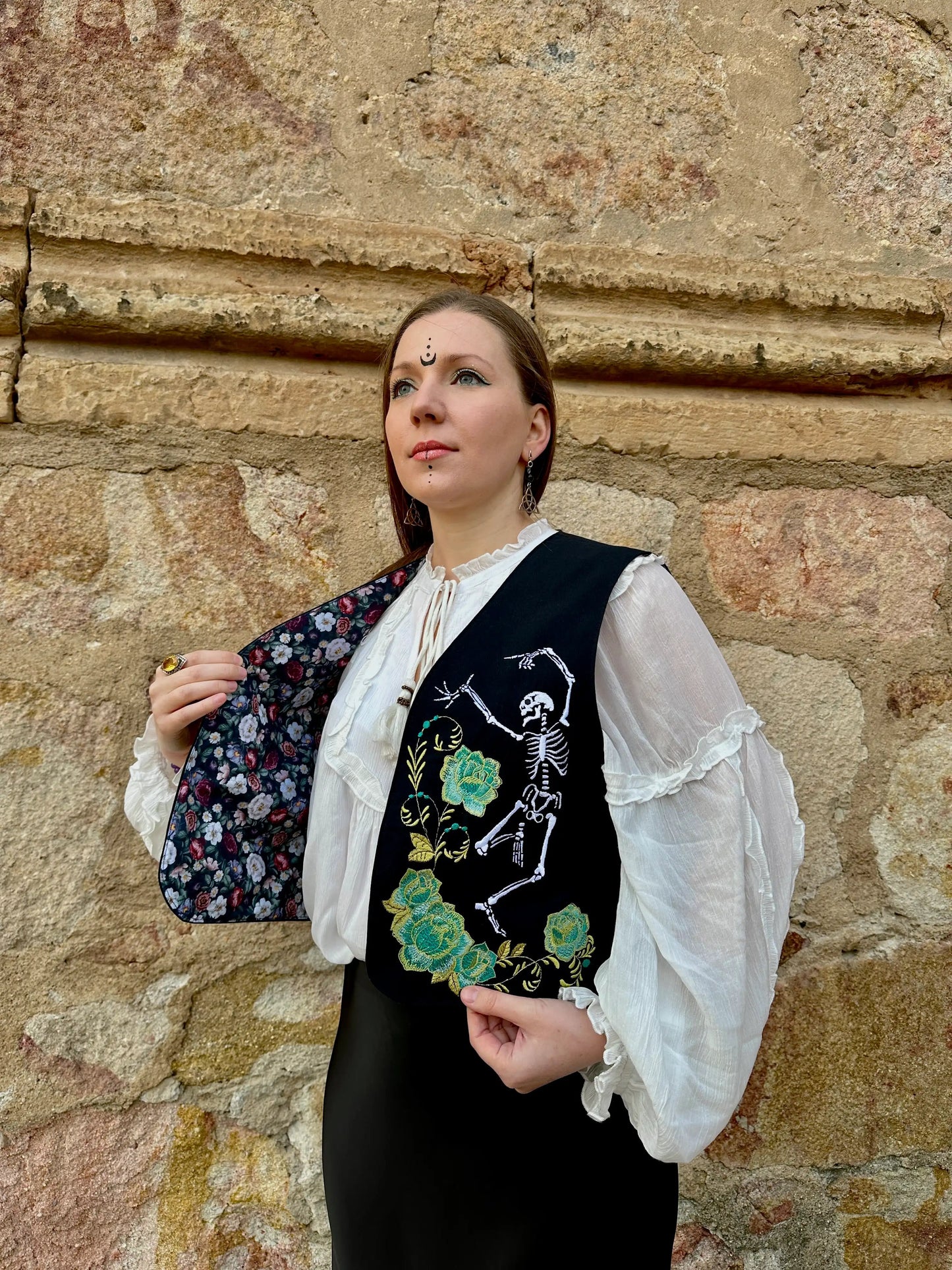 Woman wearing a black embroidered vest with floral and skeleton designs against a stone wall.