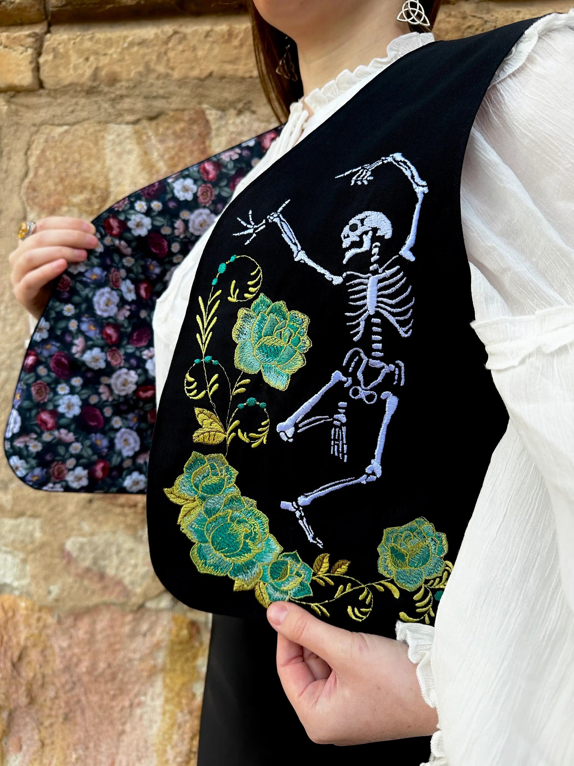 Person holding a black embroidered vest with a skeleton and floral design against a stone wall.