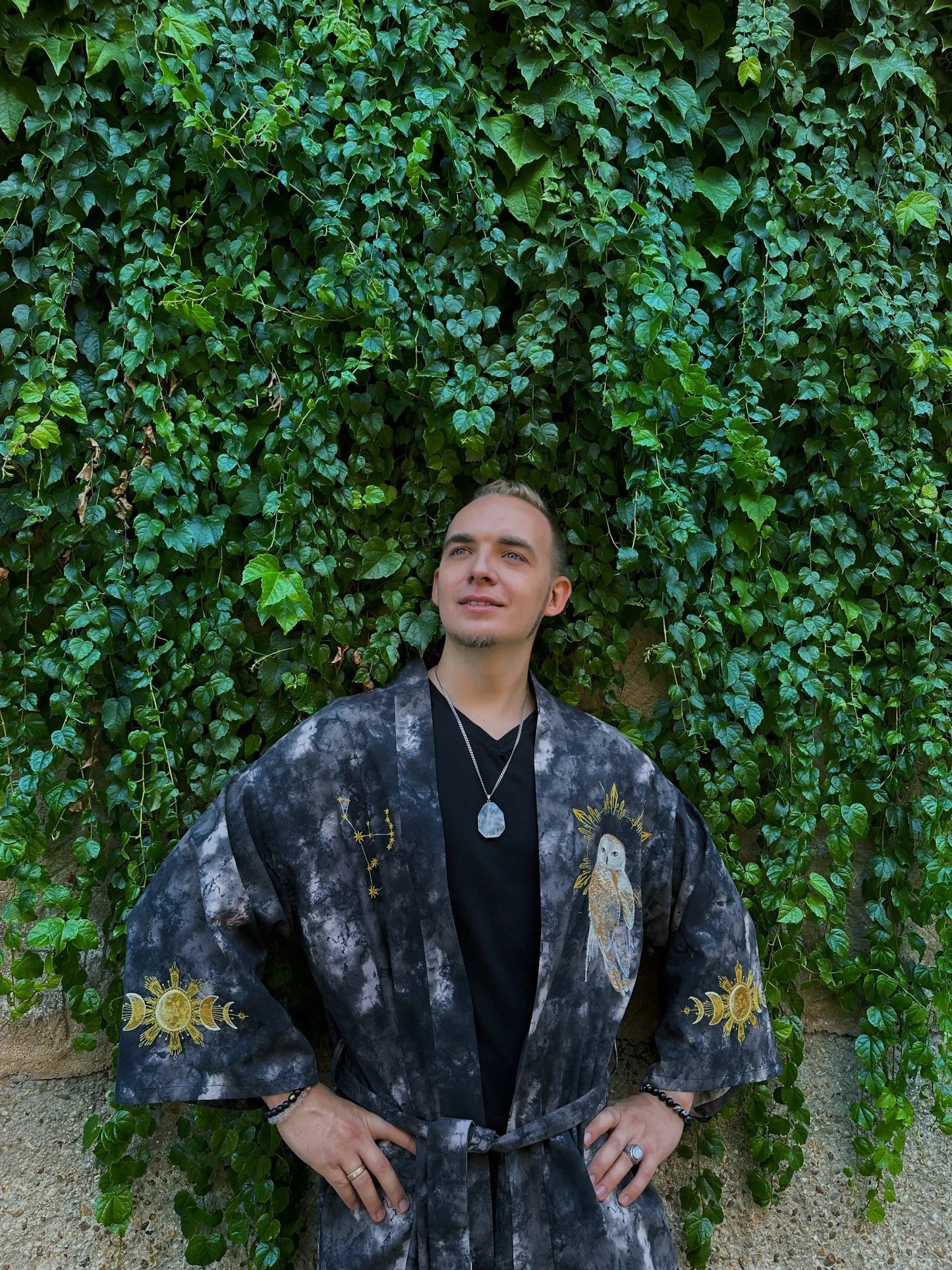 Person wearing a dark, patterned outfit standing against a green ivy-covered wall.