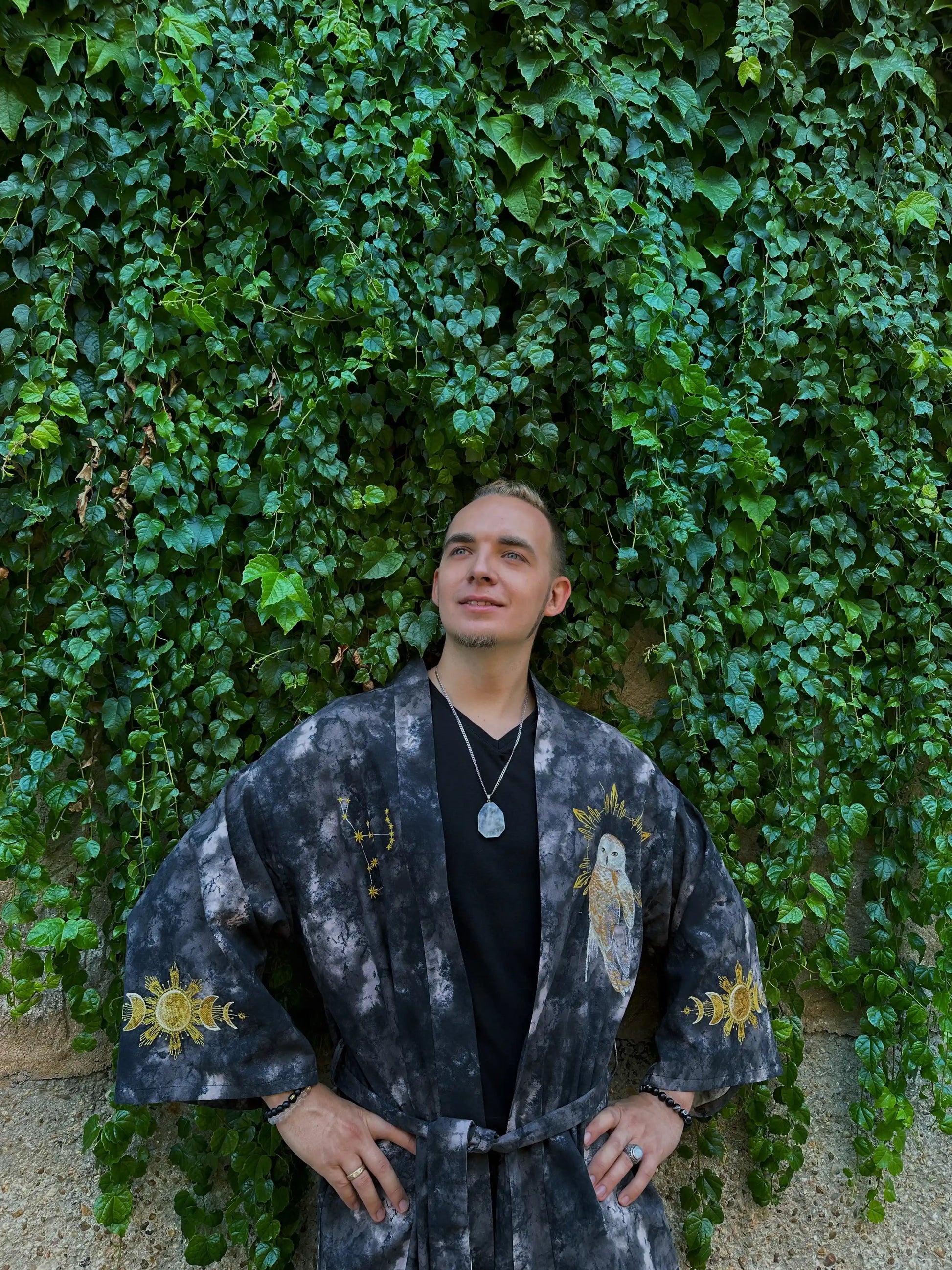 Person wearing a dark, patterned outfit standing against a green ivy-covered wall.