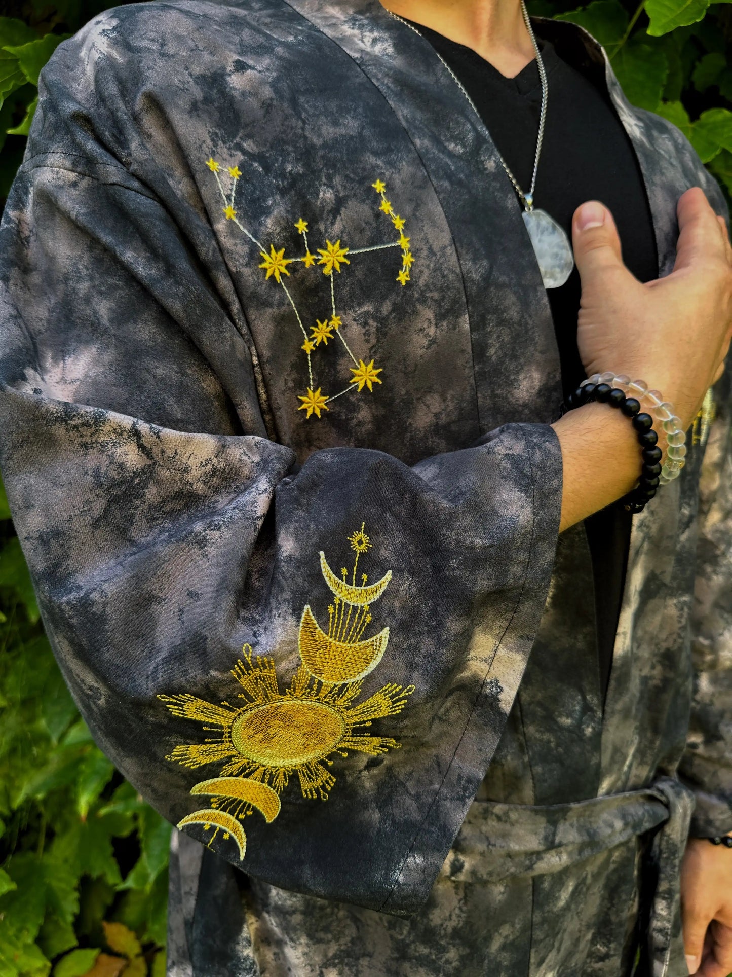 Person wearing a dark, tie-dye jacket with embroidered celestial designs on a green leafy background.