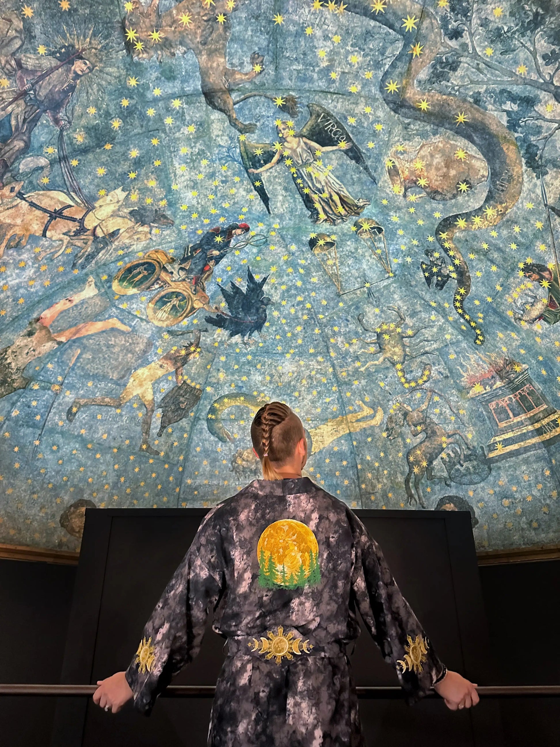 Person wearing a patterned robe in front of a decorative ceiling with celestial designs.