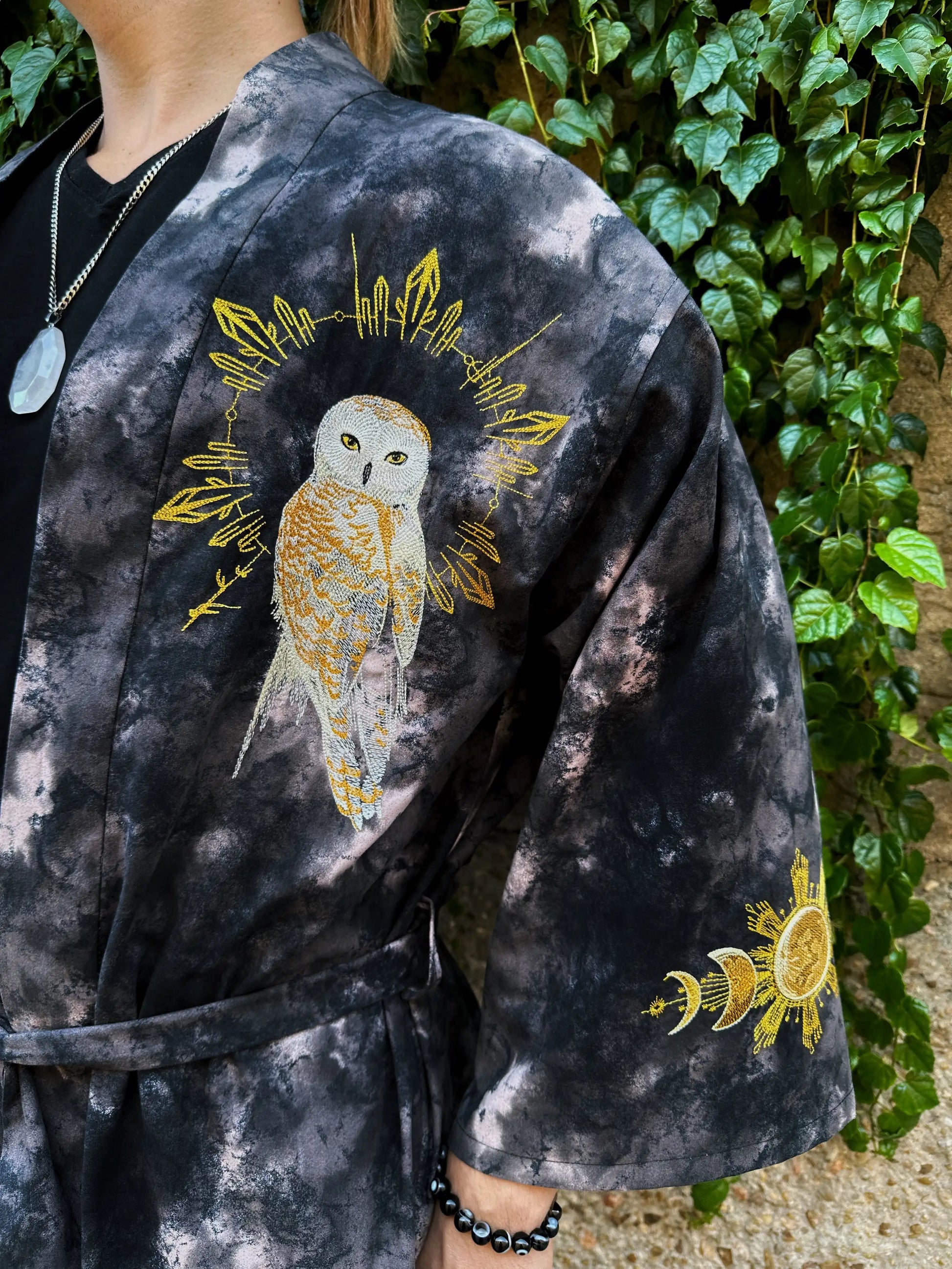 Person wearing a black tie-dye robe with gold owl and leaf designs, standing against a green leafy background.