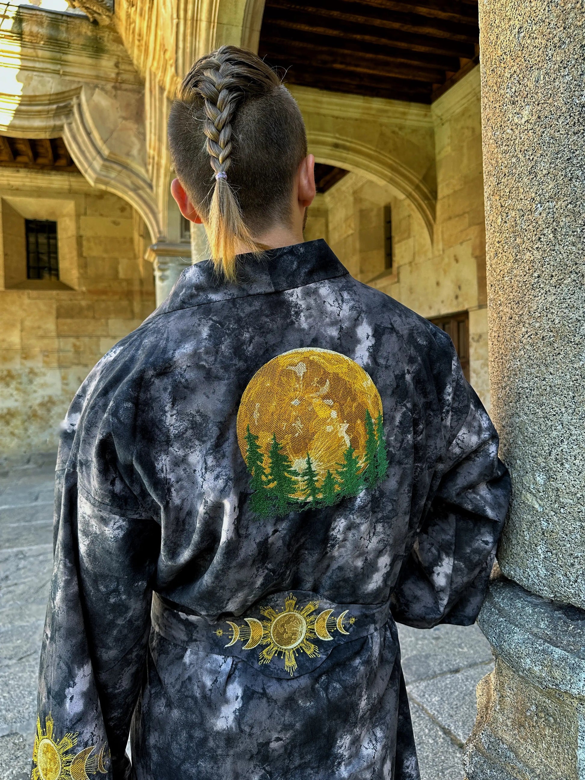 Person wearing a dark robe with a sun and tree design, standing in an architectural setting.