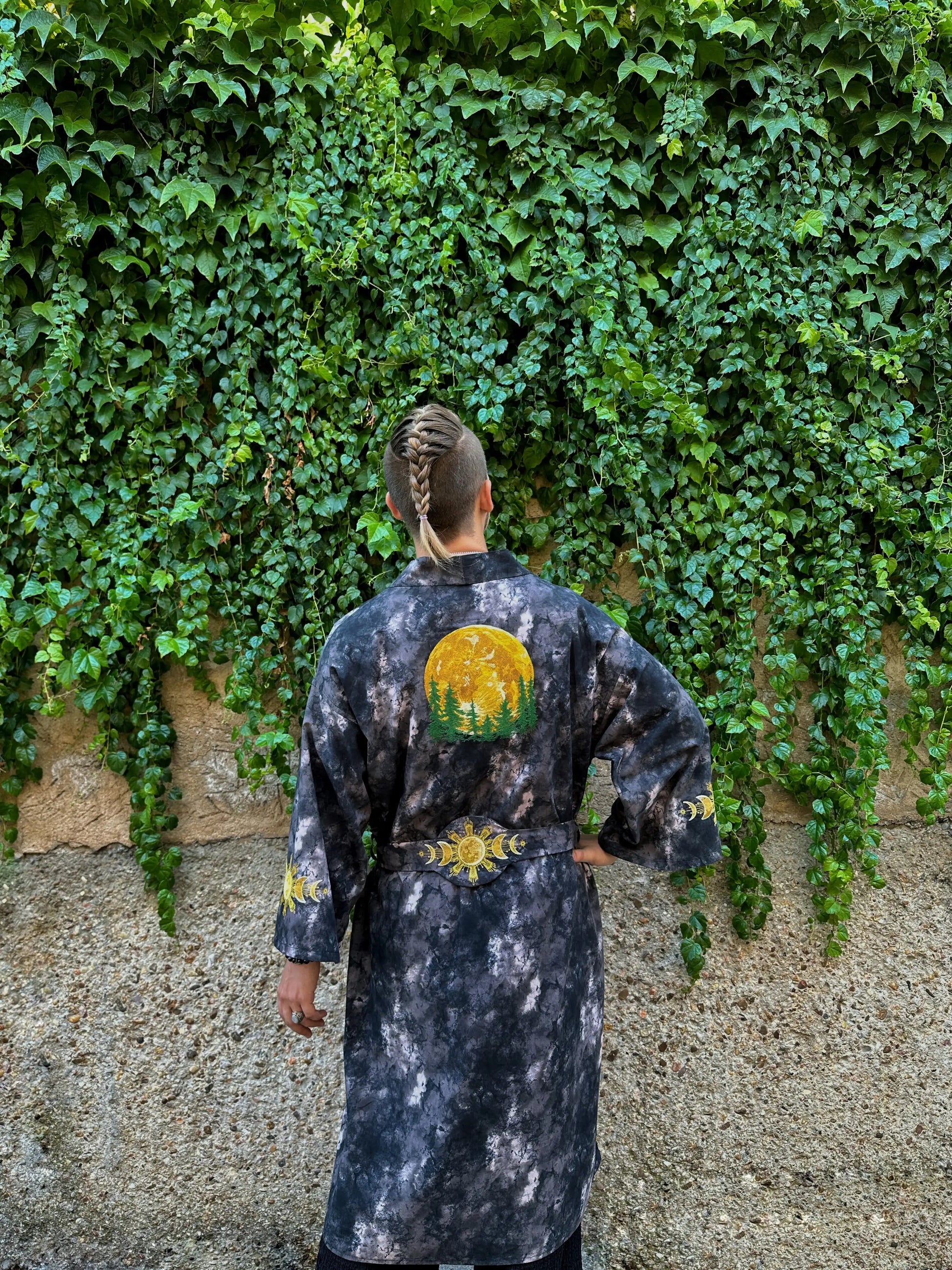 Person wearing a black tie-dye robe with a sun design, standing against a wall covered in green ivy.