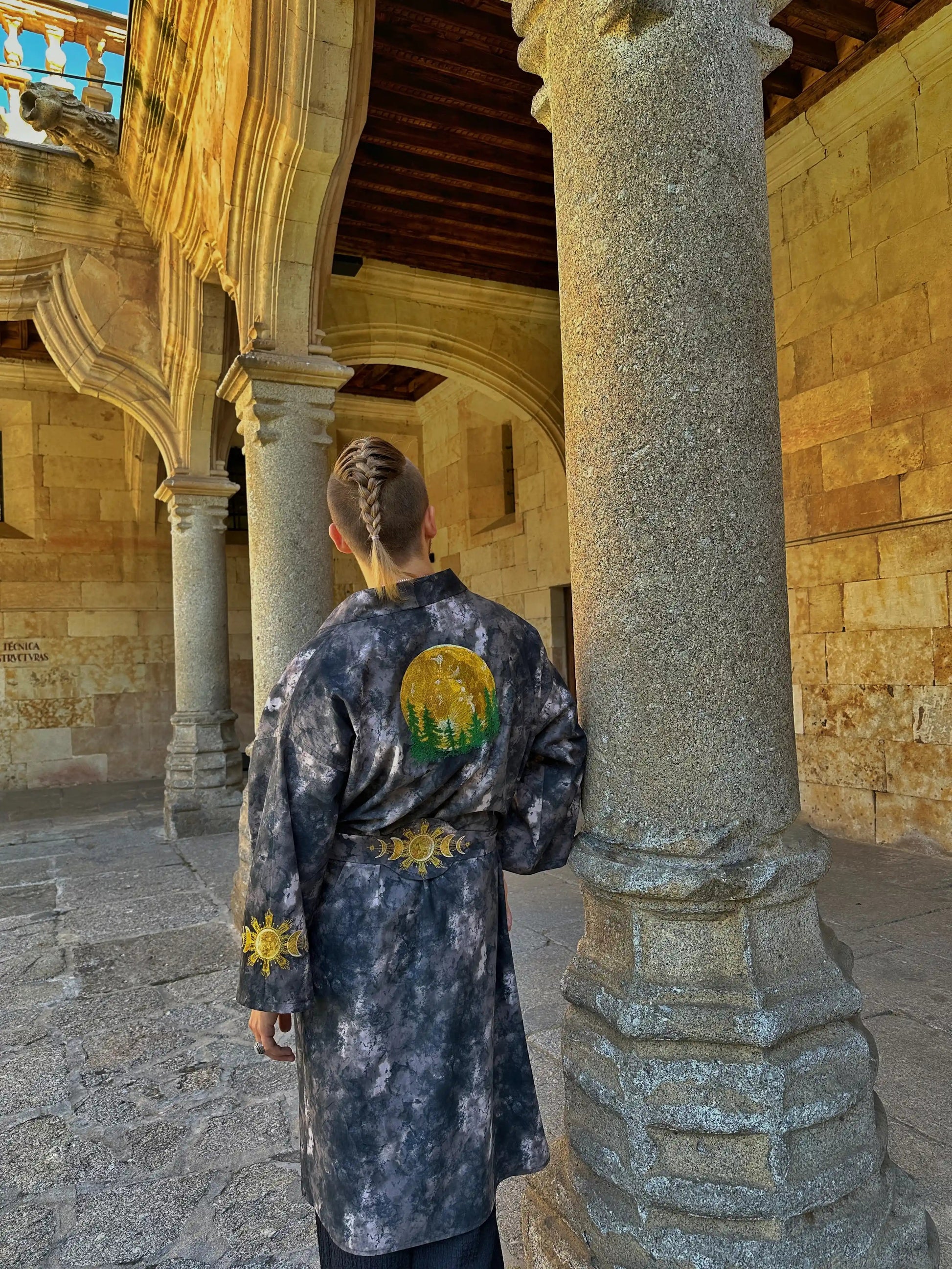 Person wearing a patterned coat with a circular design, standing in a stone archway.