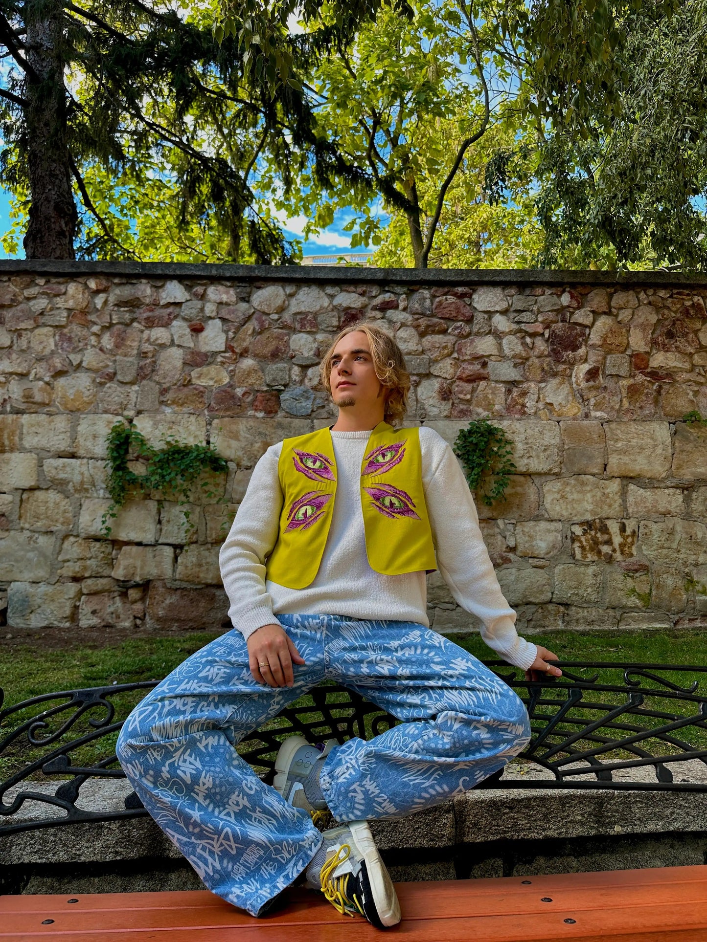 Person sitting on a bench wearing a yellow vest with eye designs and blue pants, against a stone wall and tree background.