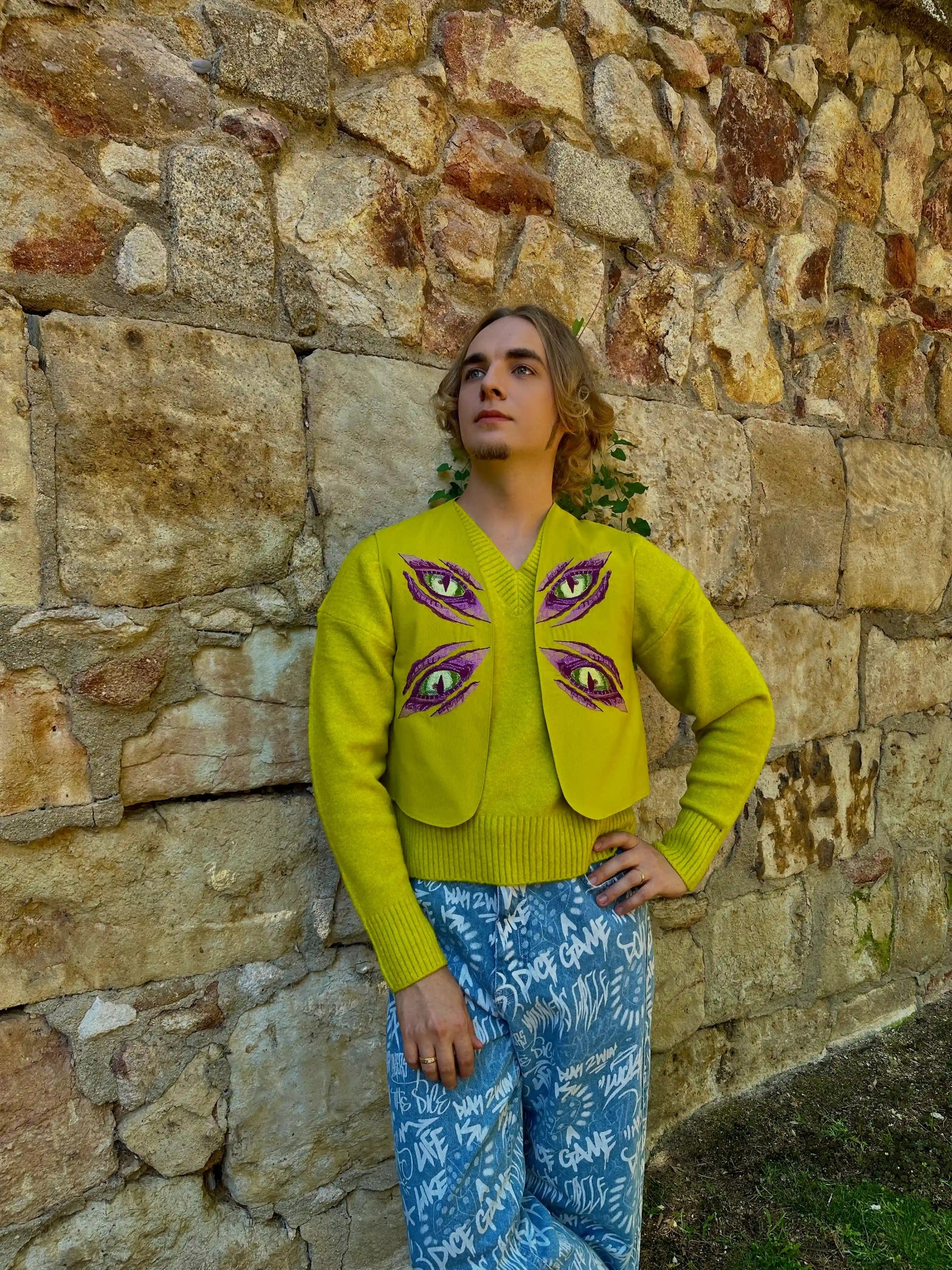 Person wearing a bright yellow cardigan with eye designs against a stone wall.
