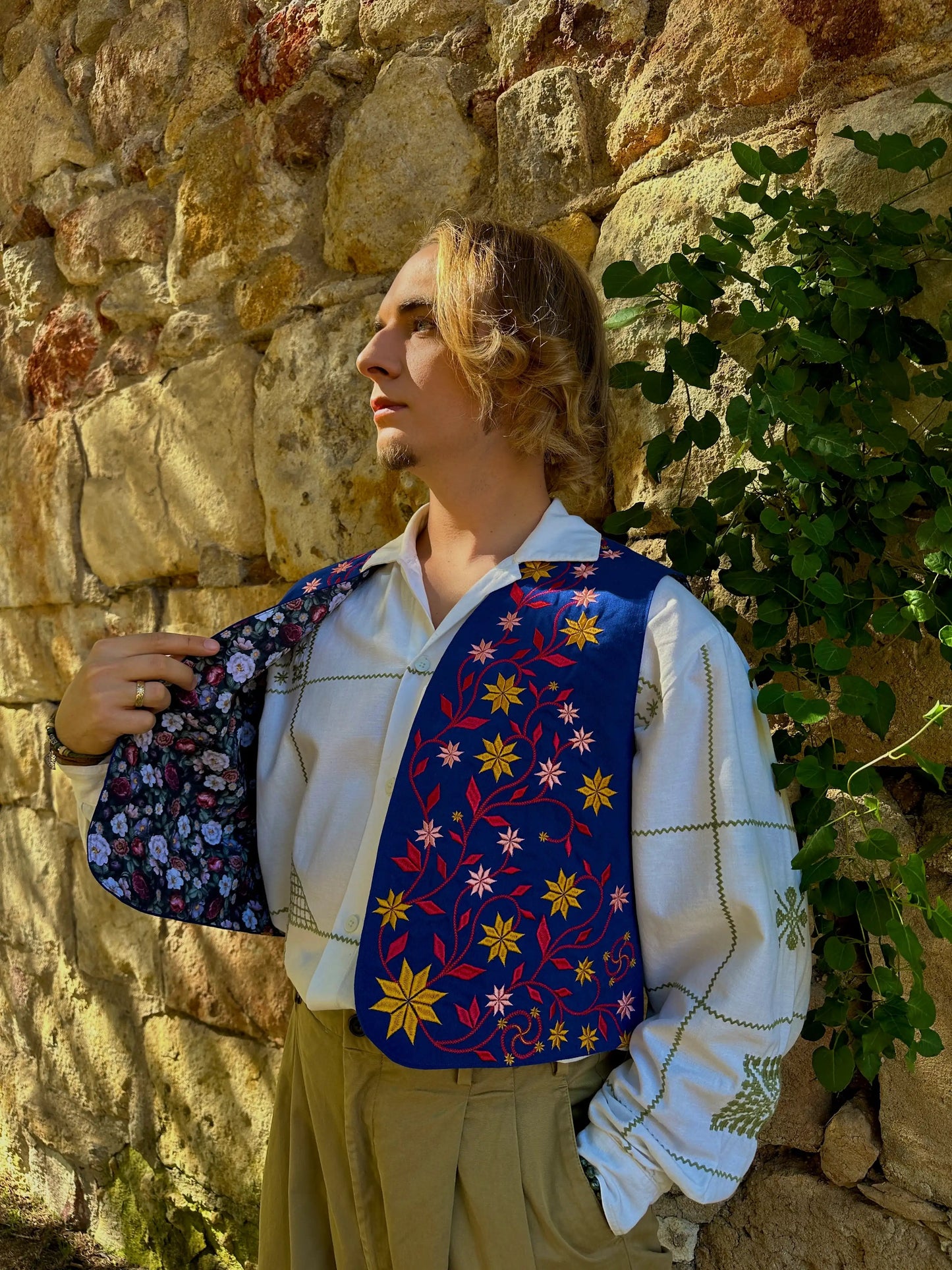 Person wearing a colorful floral-patterned vest against a stone wall with greenery.