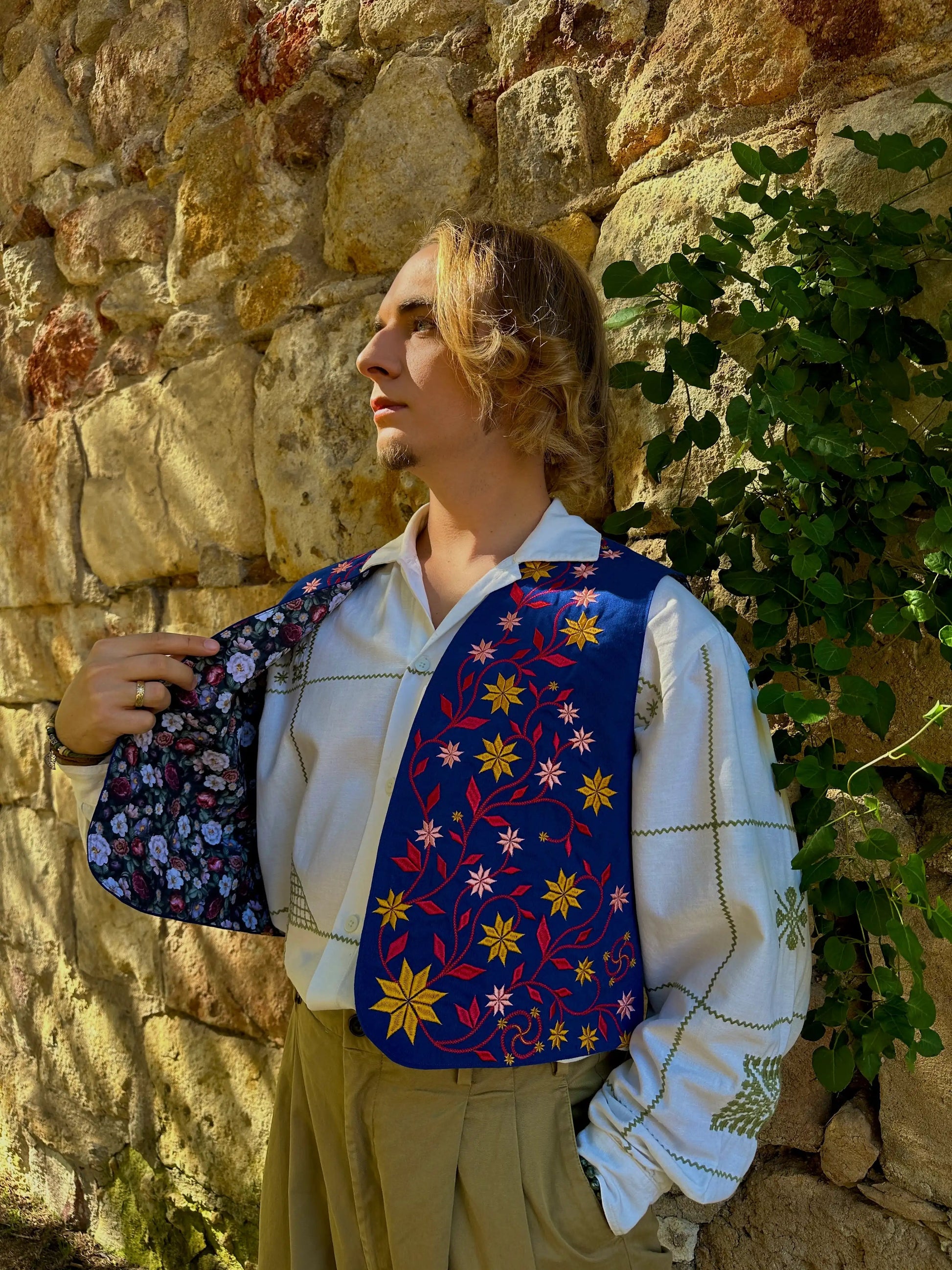 Person wearing a colorful floral-patterned vest against a stone wall with greenery.