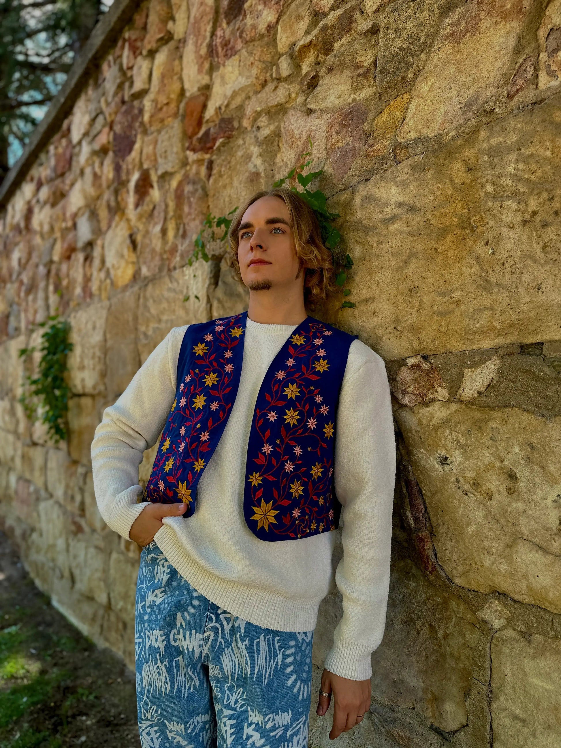 Person wearing a colorful vest and blue pants against a stone wall