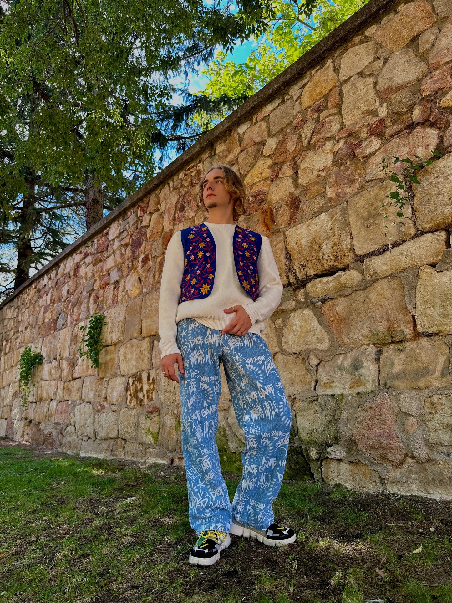 Person wearing a patterned sweater and pants standing against a stone wall with trees in the background