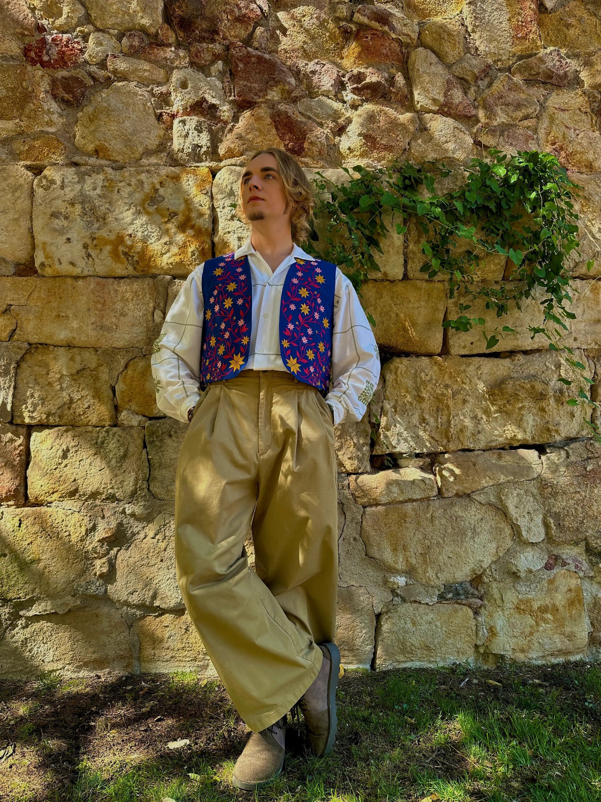 Person wearing a colorful vest and beige pants against a stone wall with greenery.