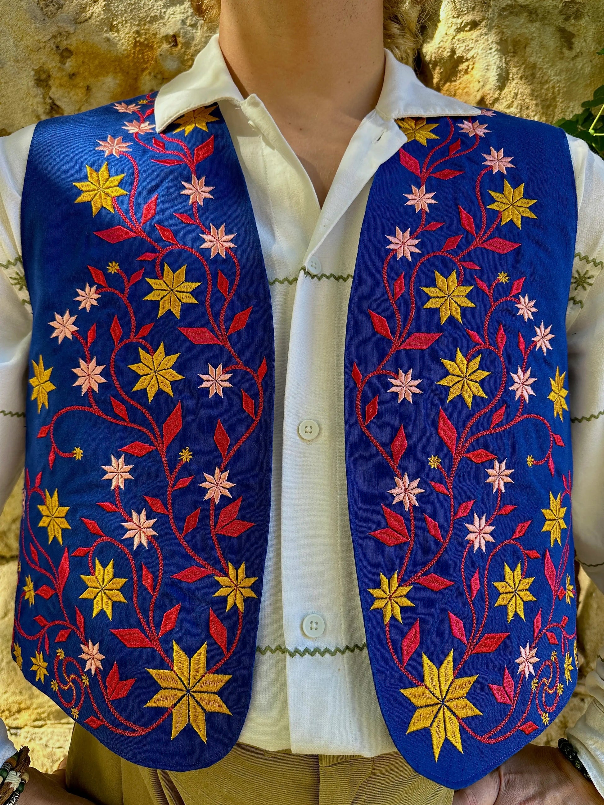 Mannequin wearing a white shirt with a colorful embroidered vest against a stone wall background
