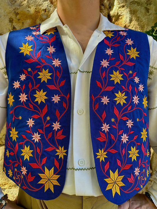 Mannequin wearing a white shirt with a colorful embroidered vest against a stone wall background