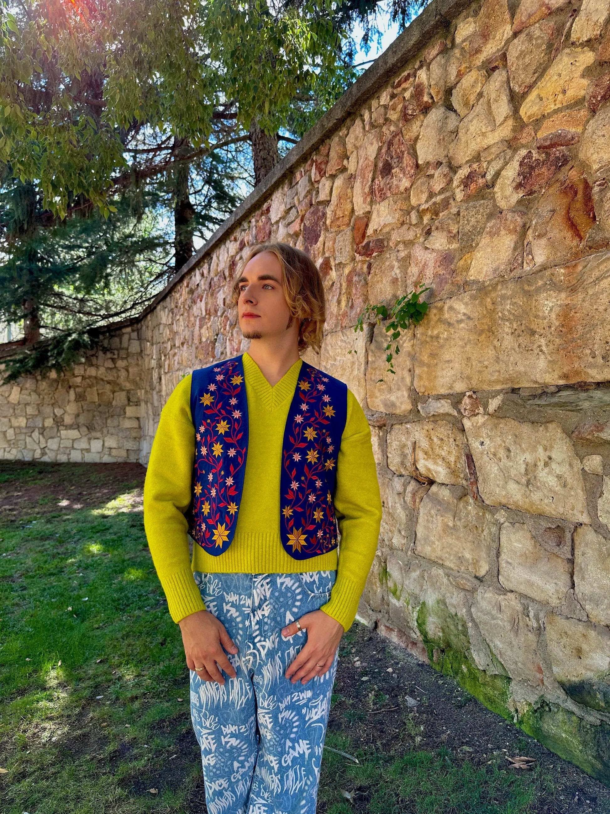Person wearing a bright yellow sweater and colorful vest standing against a stone wall with trees in the background
