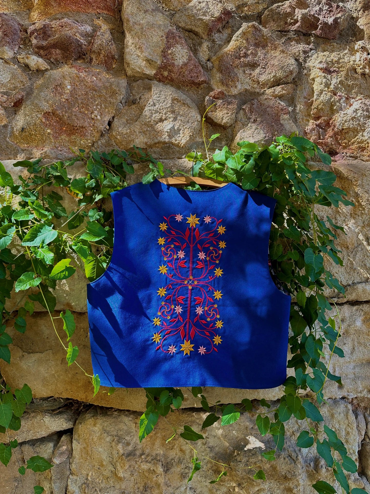Blue embroidered vest hanging against a stone wall with greenery