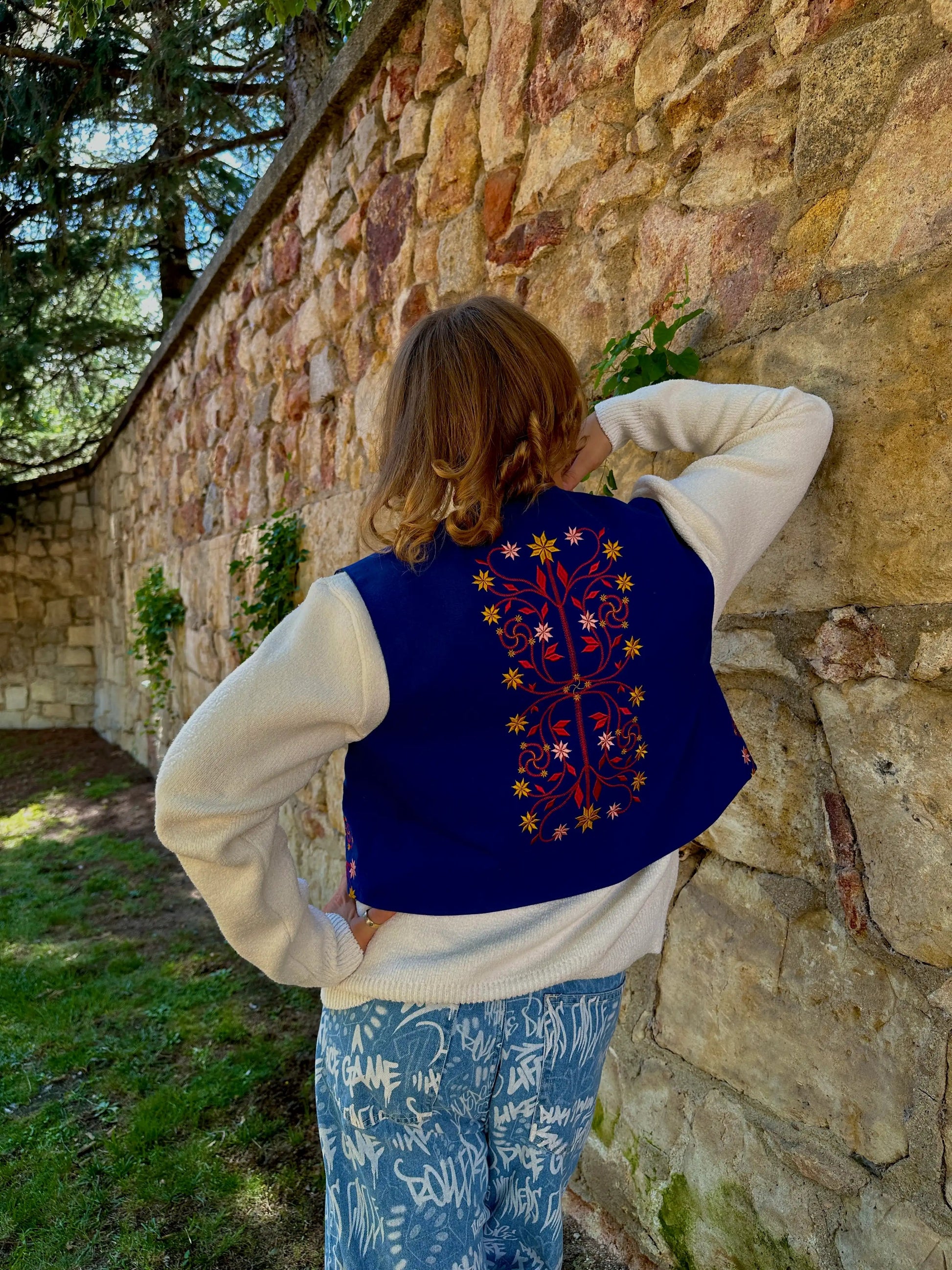Person wearing a blue vest with floral embroidery, standing against a stone wall.