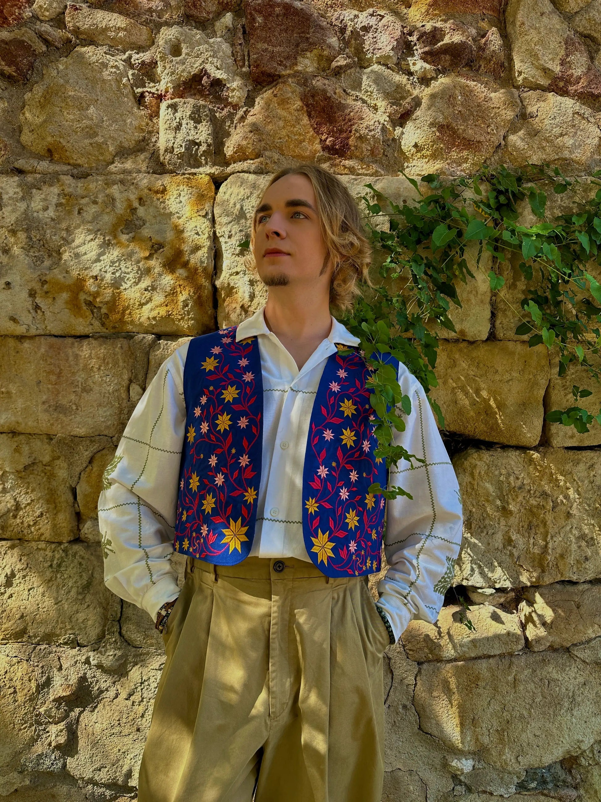 Person wearing a colorful vest and beige pants against a stone wall with greenery.