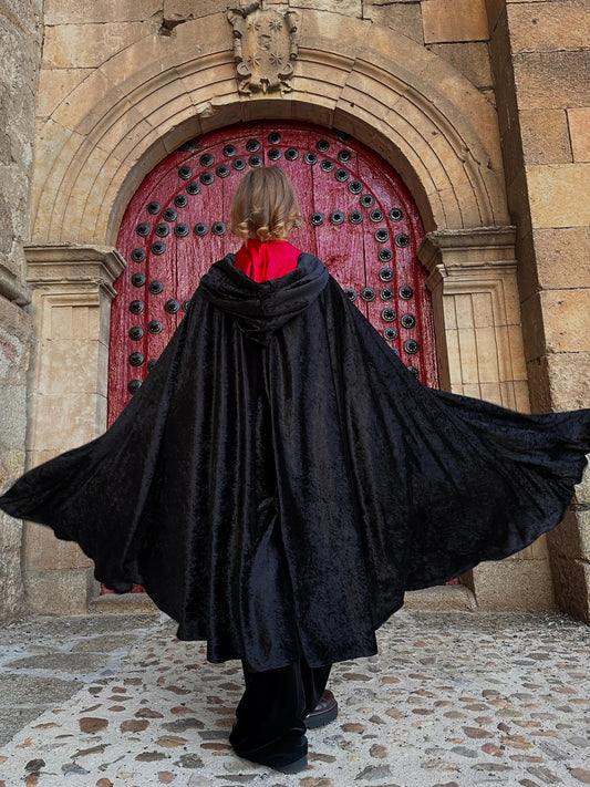 Person in a black cloak standing in front of a red door with stone architecture