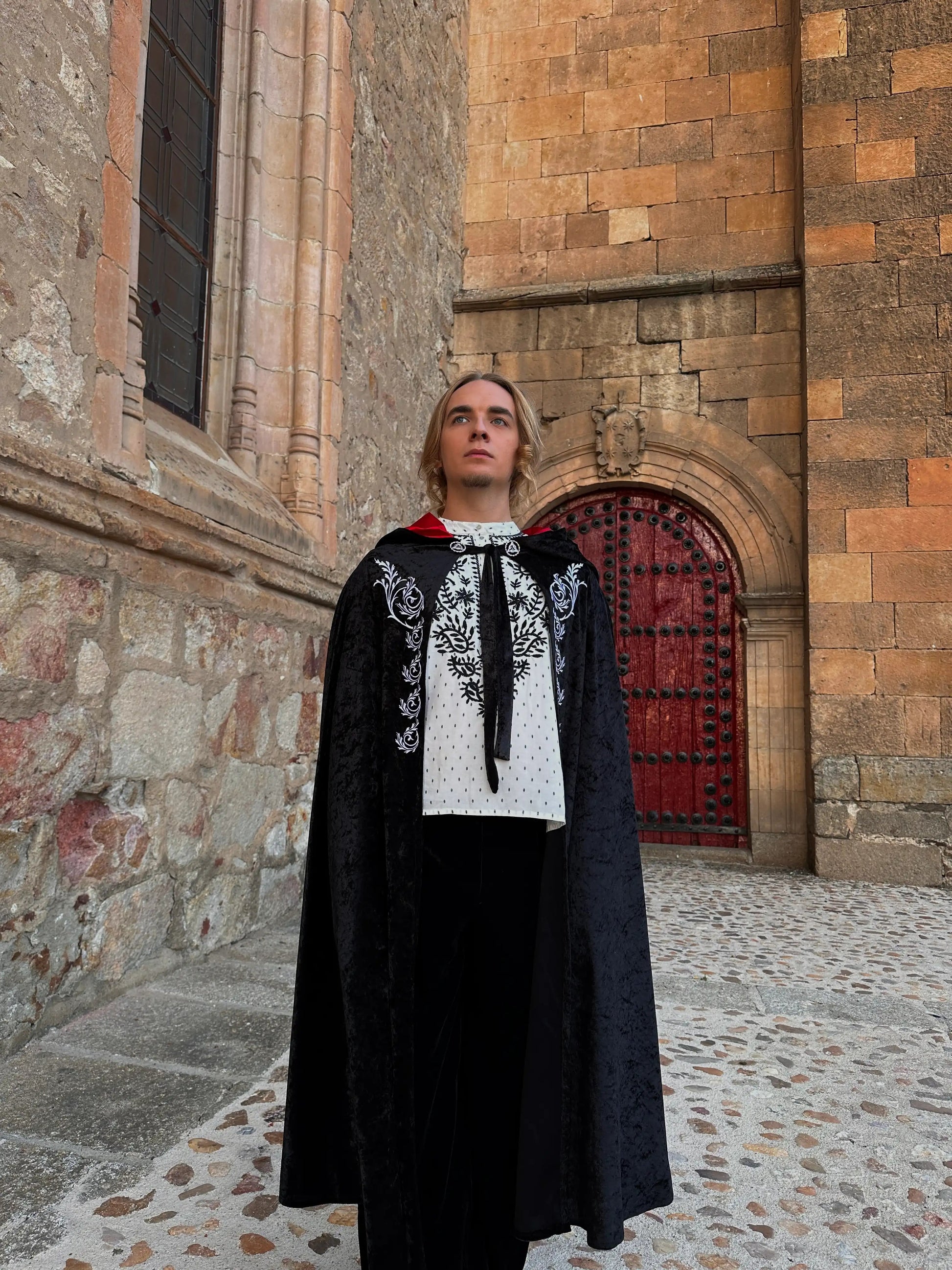 Person wearing a black cape with white embroidery standing in front of a stone building.