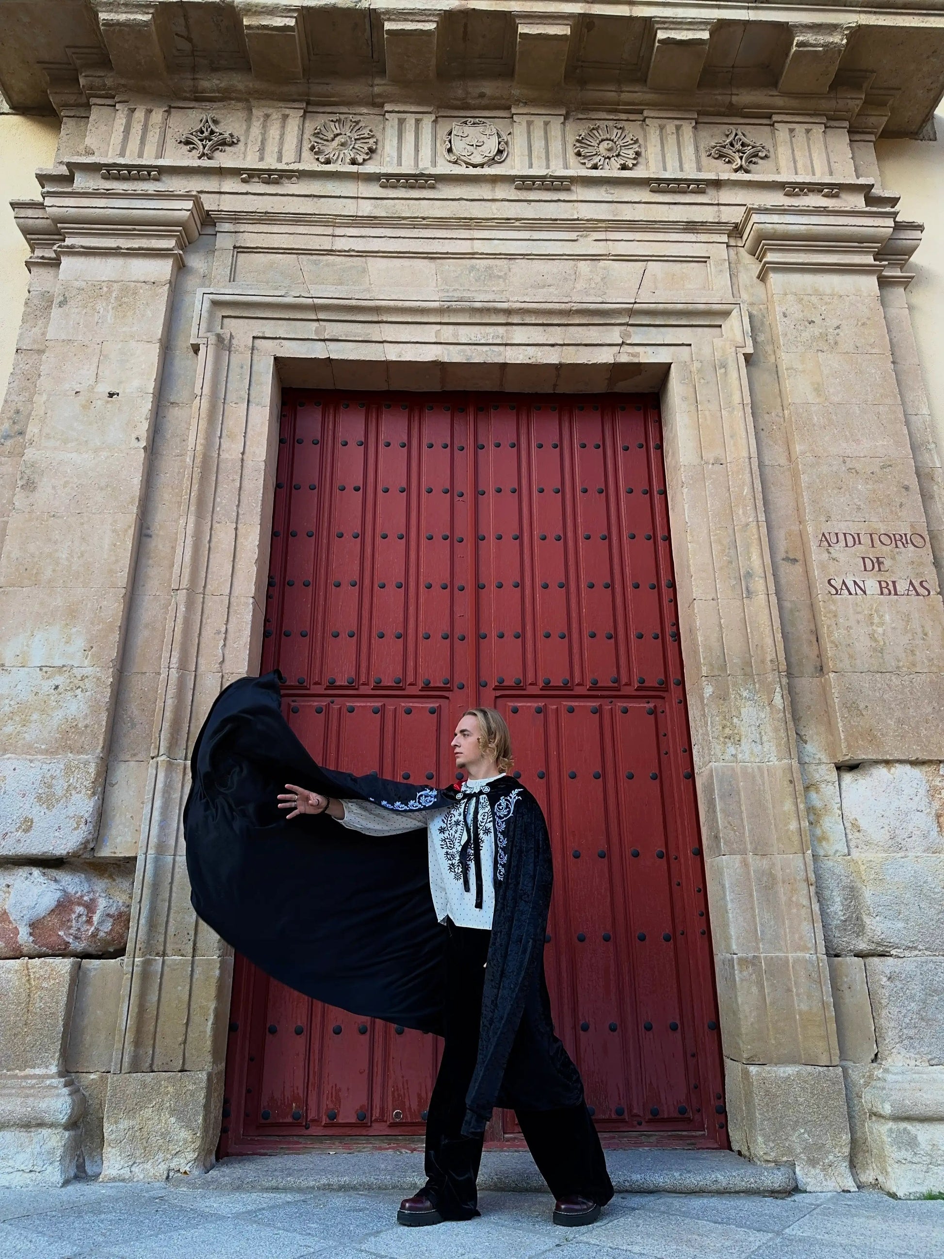 Person in formal attire posing dramatically in front of a large red door with architectural details.