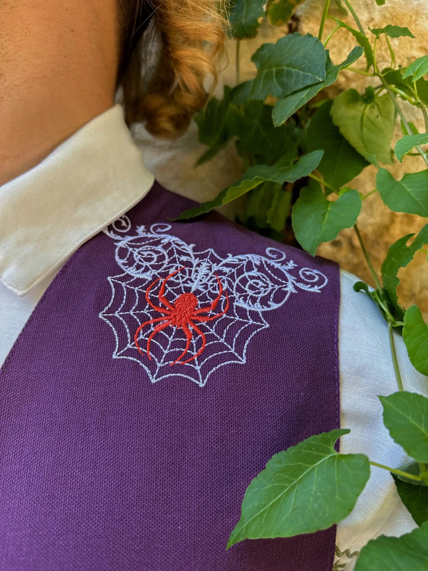 Purple garment with embroidered spider and web design, surrounded by green leaves.
