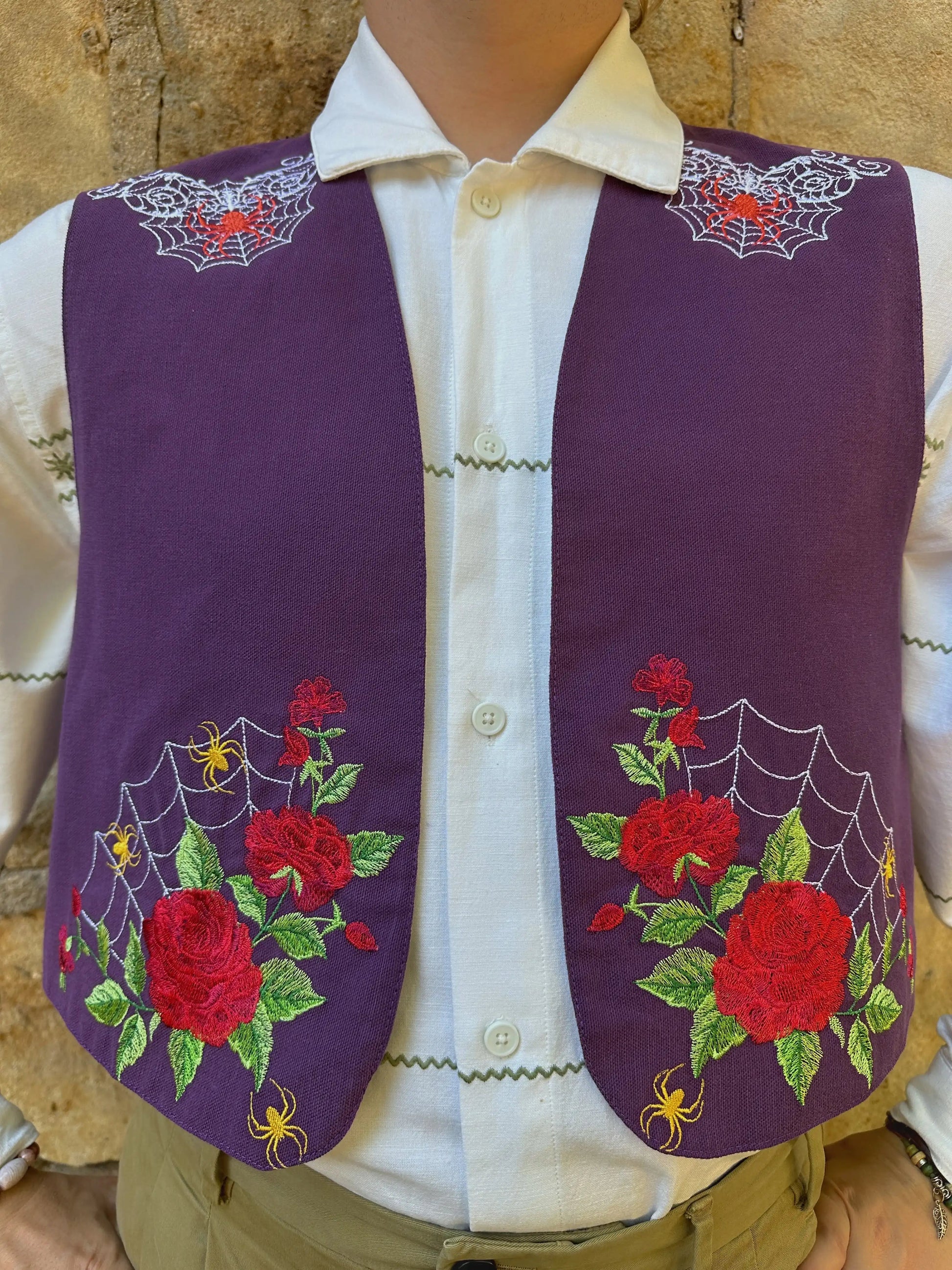 Purple embroidered vest with red roses and green leaves on a mannequin against a stone wall background.