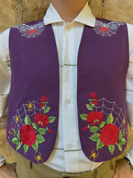 Purple embroidered vest with red roses and green leaves on a mannequin against a stone wall background.