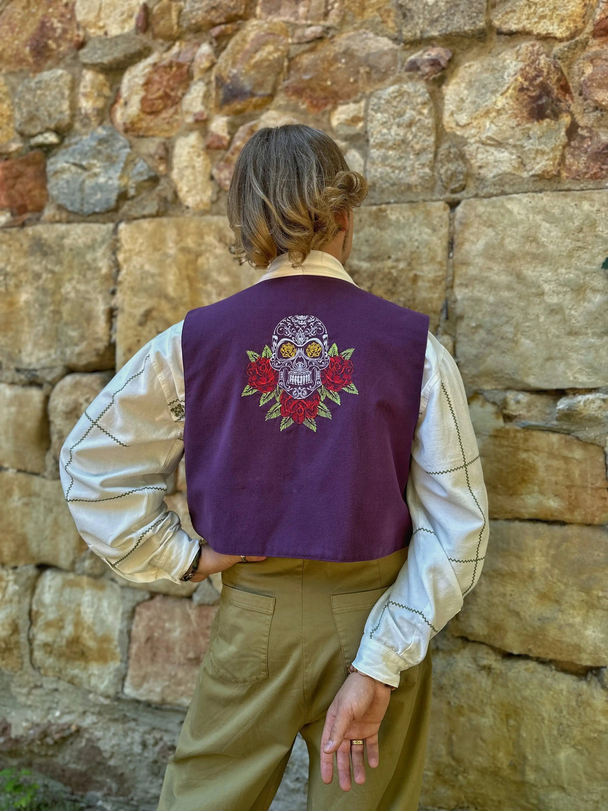 Person wearing a purple vest with a colorful skull design against a stone wall.