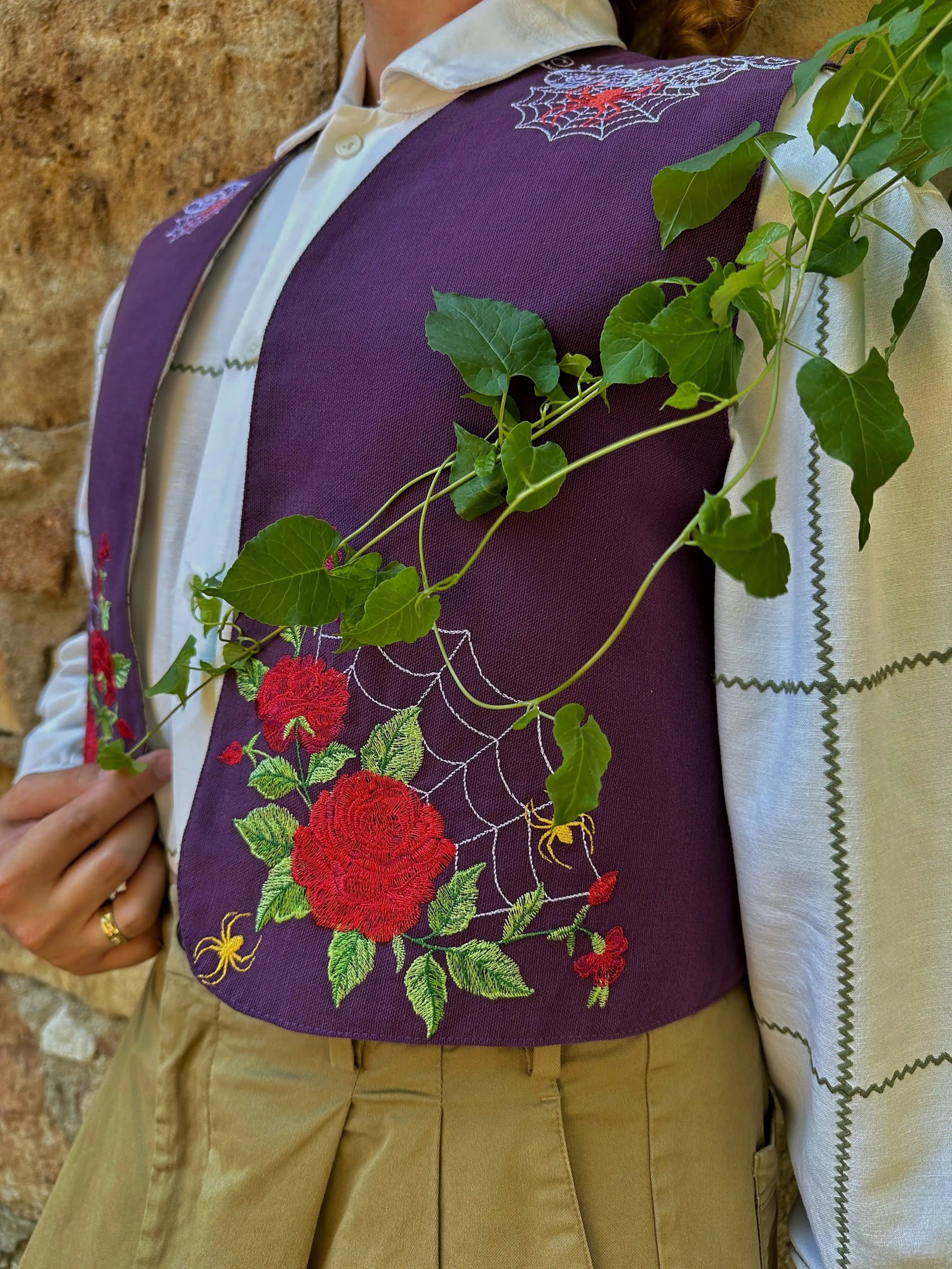 Person wearing a purple embroidered jacket with red roses and green leaves, standing against a stone wall.