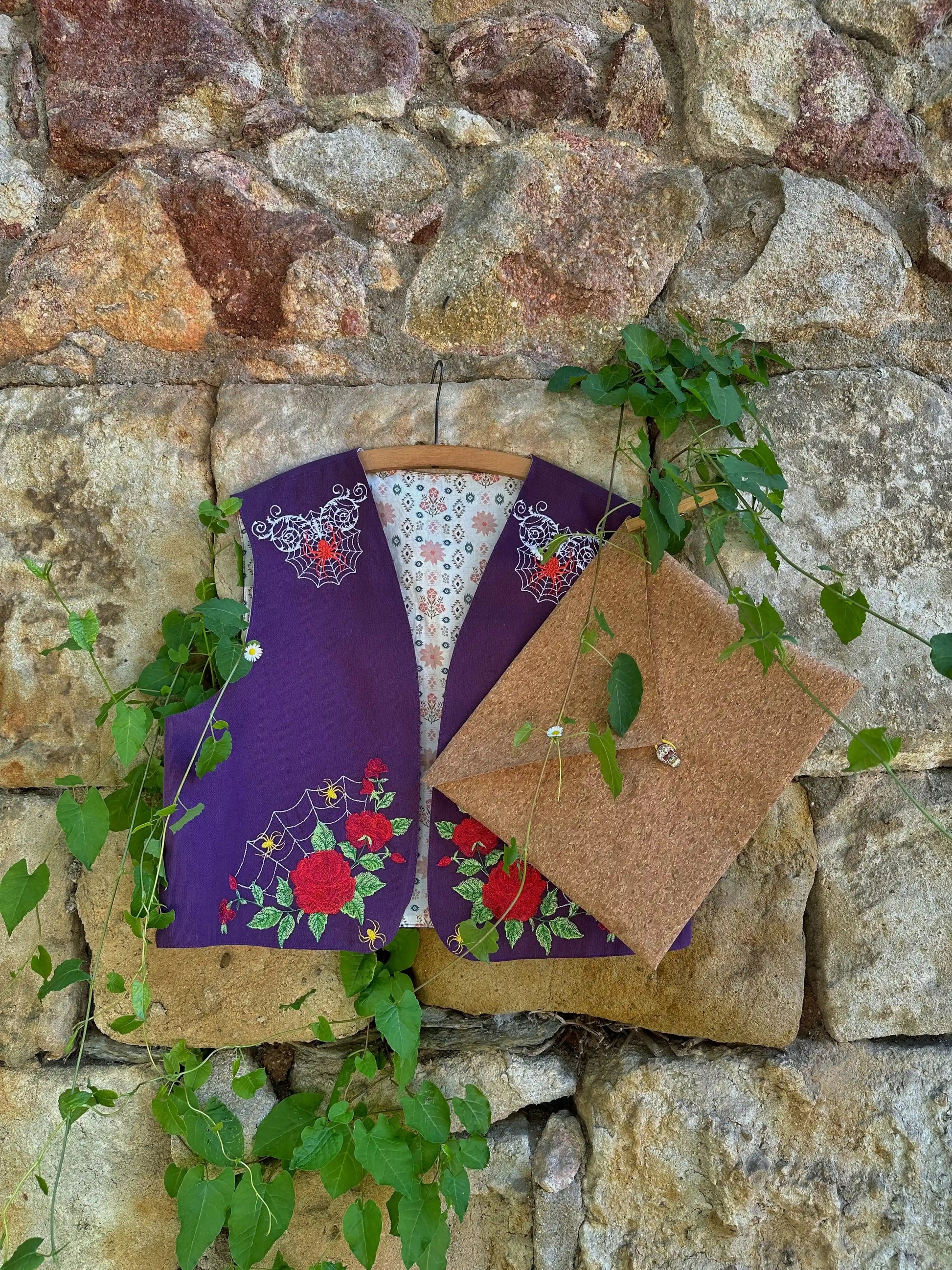 Two vests, one purple with floral patterns and the other brown, hanging against a stone wall with greenery.
