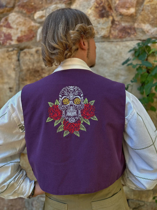 Person wearing a purple vest with a skull and roses design against a stone wall.